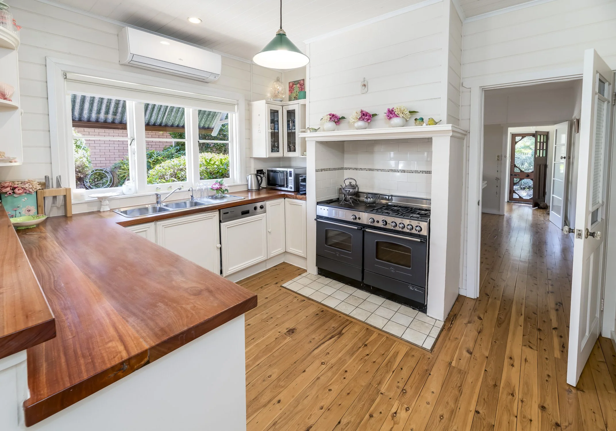 Kitchen with wooden countertops, white cabinets, and a black stove. A window above the sink shows greenery outside. There are small kitchen appliances on the counter, and decorative flowers on the mantle above the stove. Hardwood floors and a doorway