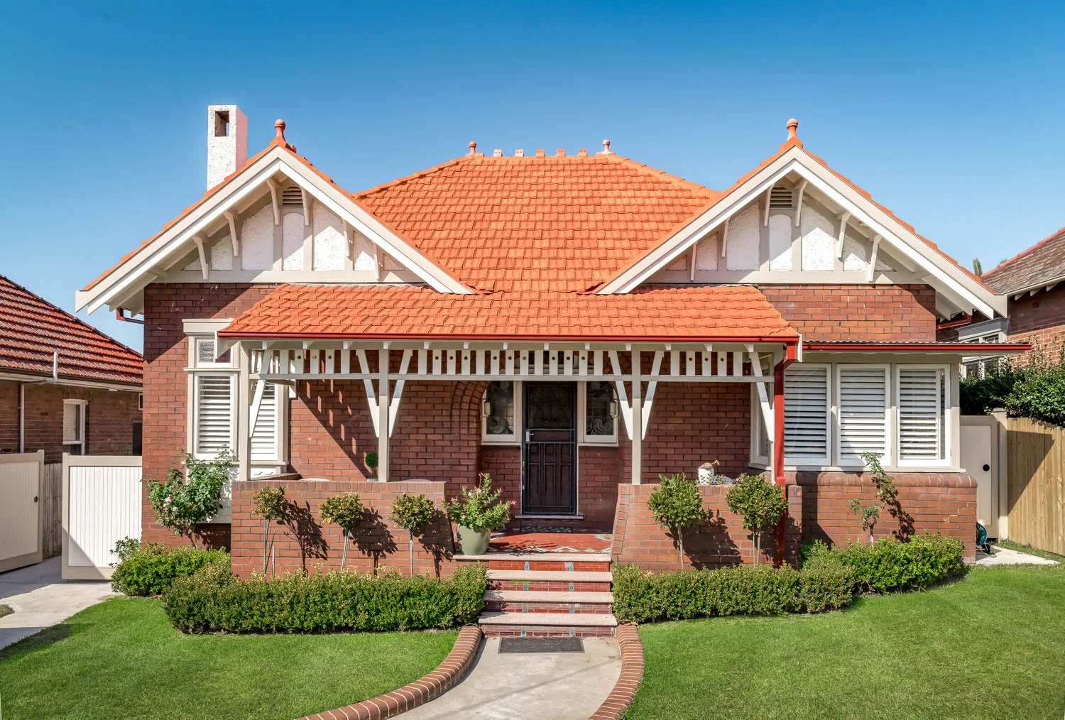 A two-story brick house with a red tiled roof, white decorative trim, and a front porch with white support beams. There are stairs leading to the black front door, with neatly trimmed bushes and small trees in the front yard.