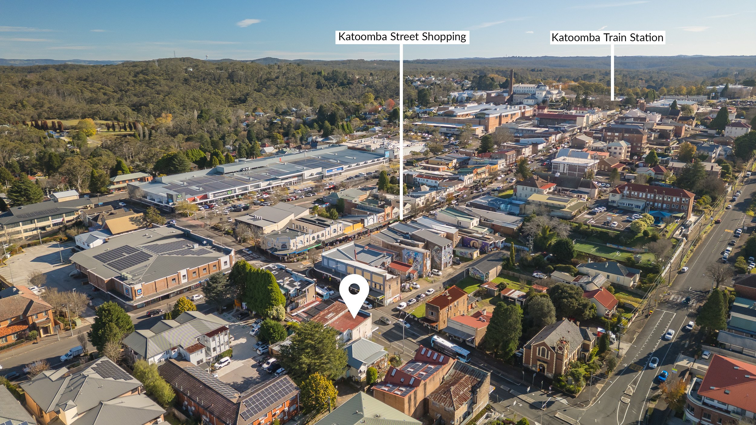 Aerial view of Katoomba town showing shopping center, train station, surrounding buildings, parking lots, and nearby green hills under a clear blue sky.