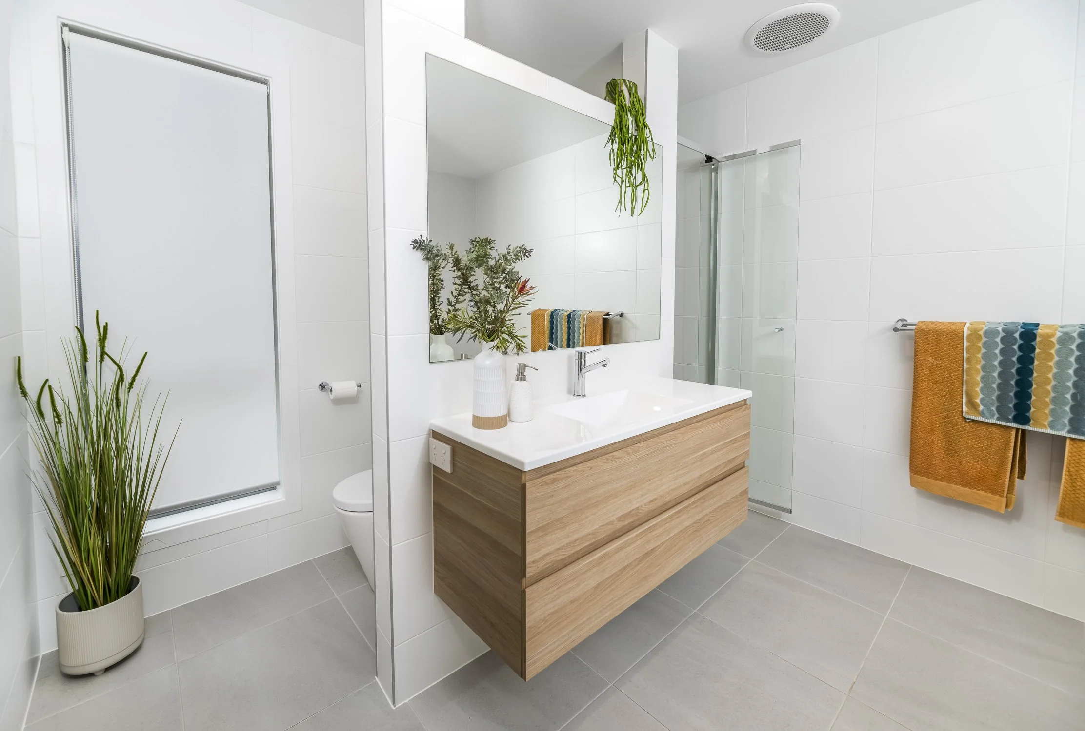 Modern bathroom with a white tiled wall, a wooden vanity with a white countertop, a large mirror, towels, a potted plant, and a shower with a glass door.