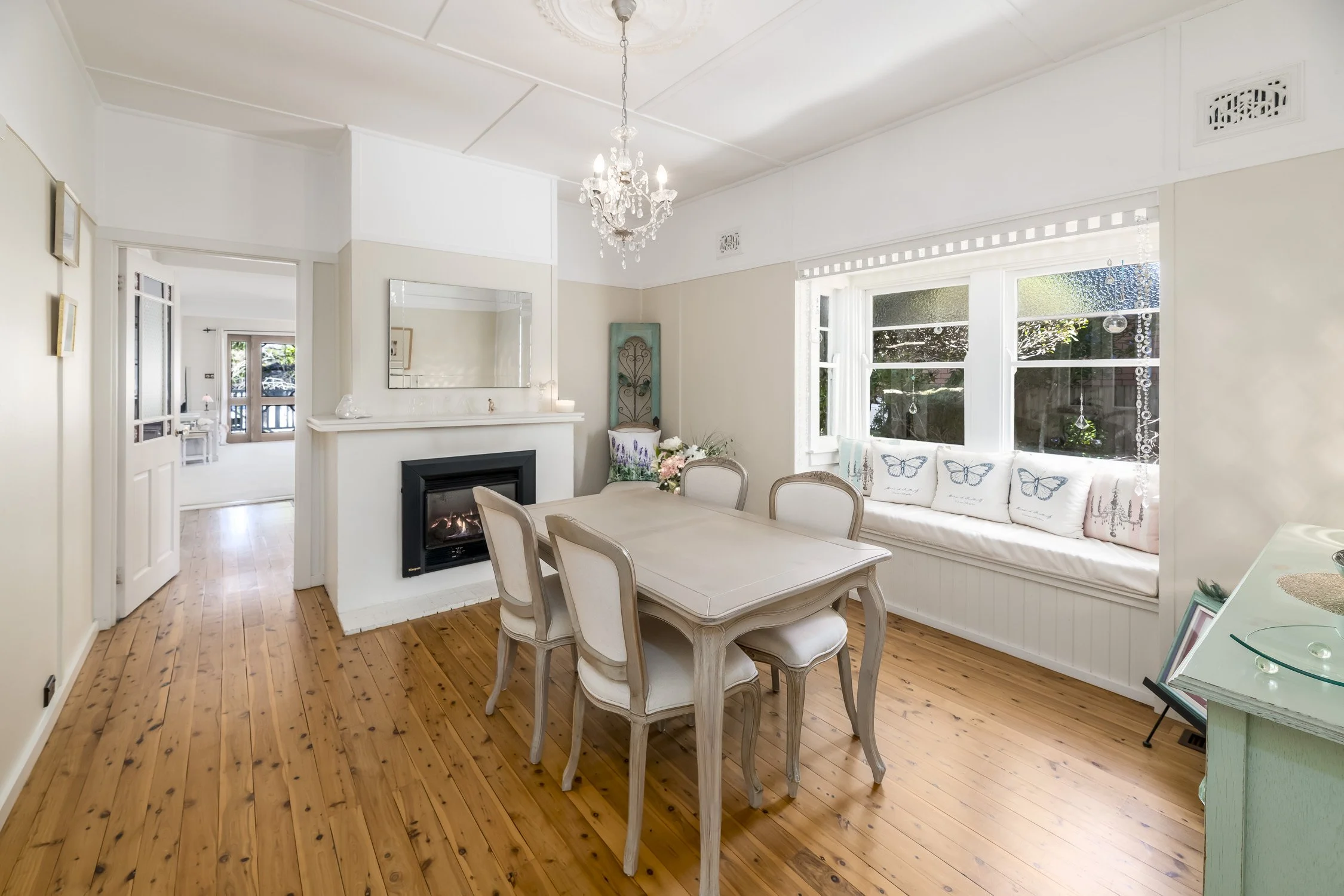 Bright dining room with a wooden table, white upholstered chairs, a wall with large windows and butterfly pillows on a window seat, and a fireplace with a mirror above it.