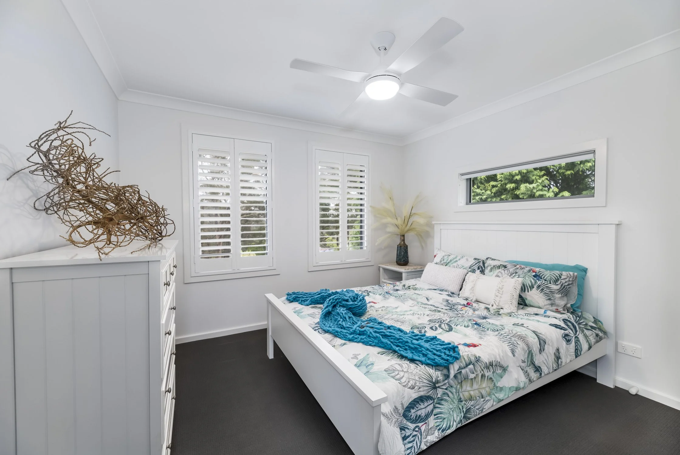 A bright bedroom with white walls, a white bed with tropical-themed bedding, and a bedside table with a tall vase of decorative pampas grass. There is a white dresser with a large driftwood sculpture, and three windows with white shutters and a horiz