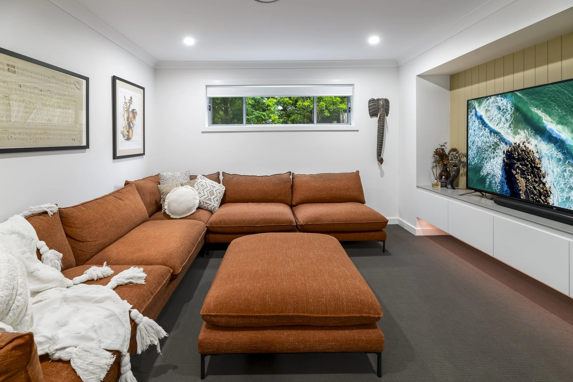 Living room with white walls, orange sectional sofa, throw pillows, wall art, window with greenery outside, elephant sculpture on wall, large television with ocean image, and modern decor.