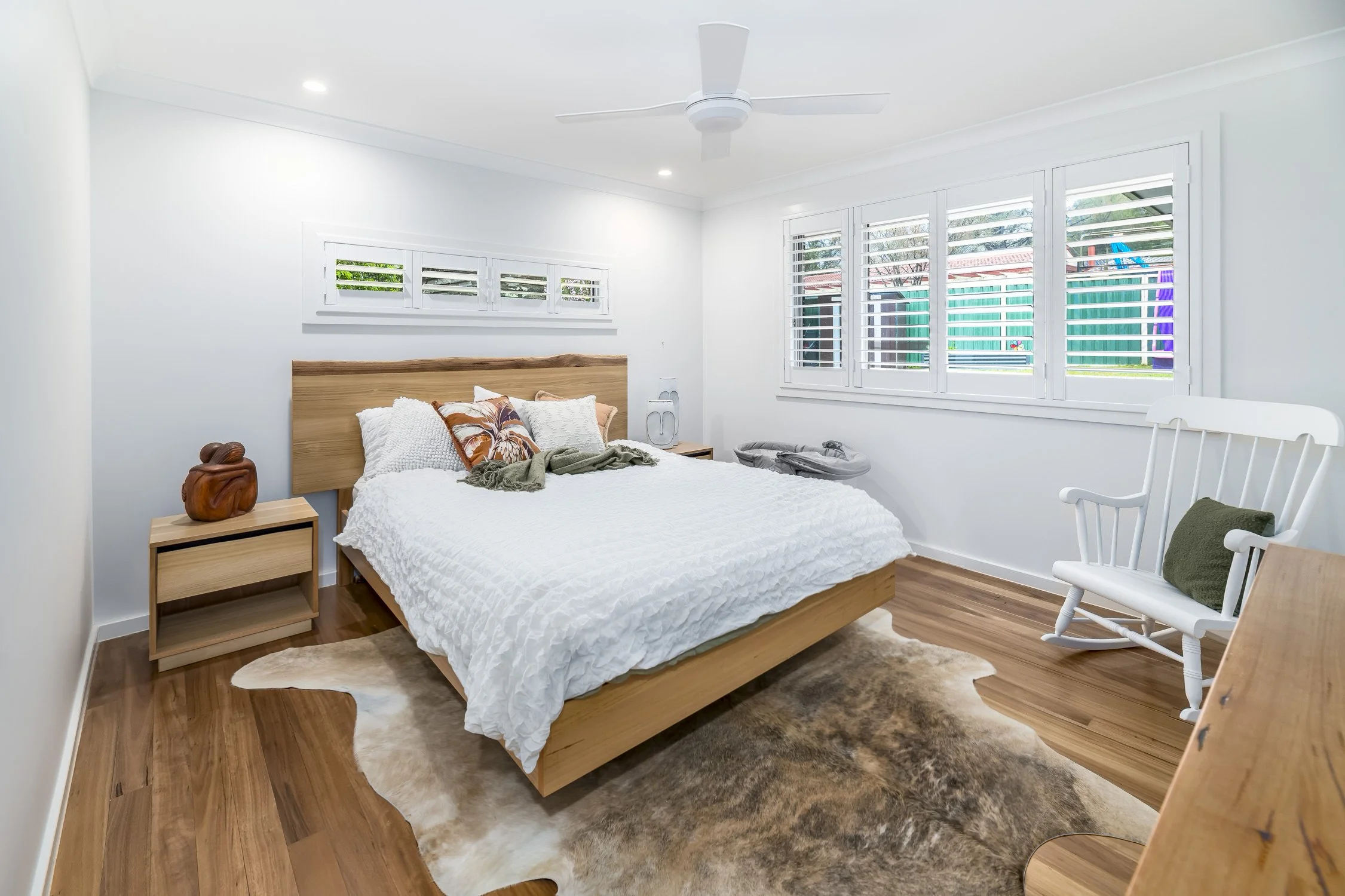 A bedroom with white walls, wooden furniture, a white bed with pillows, a window with shutters, a white rocking chair, and a cowhide rug on wooden floor.