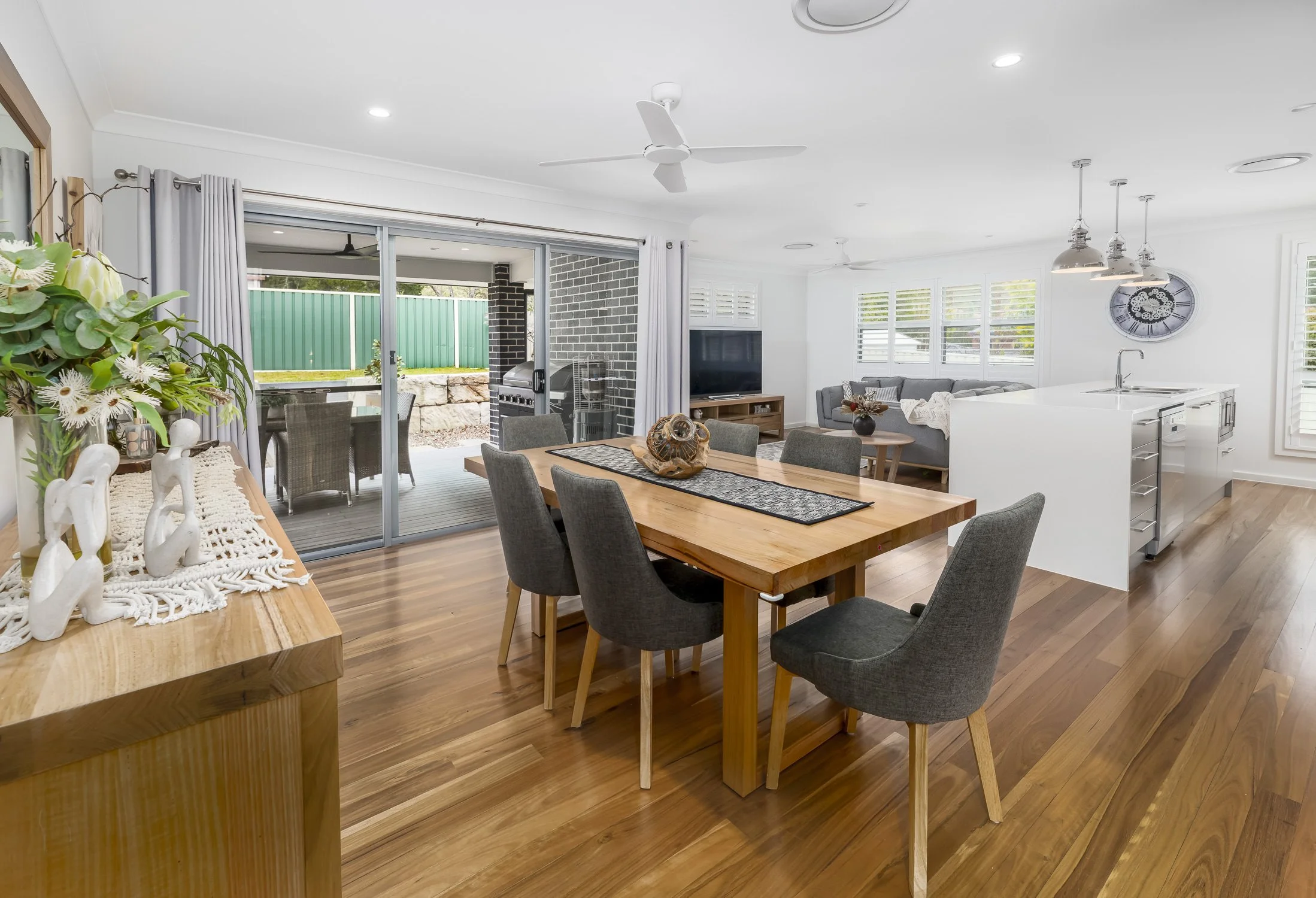 Open concept dining and living room with hardwood floors, a wooden dining table with six dark gray chairs, a white kitchen island, and a sliding glass door leading to an outdoor patio. There are white walls, large windows with blinds, and a ceiling f