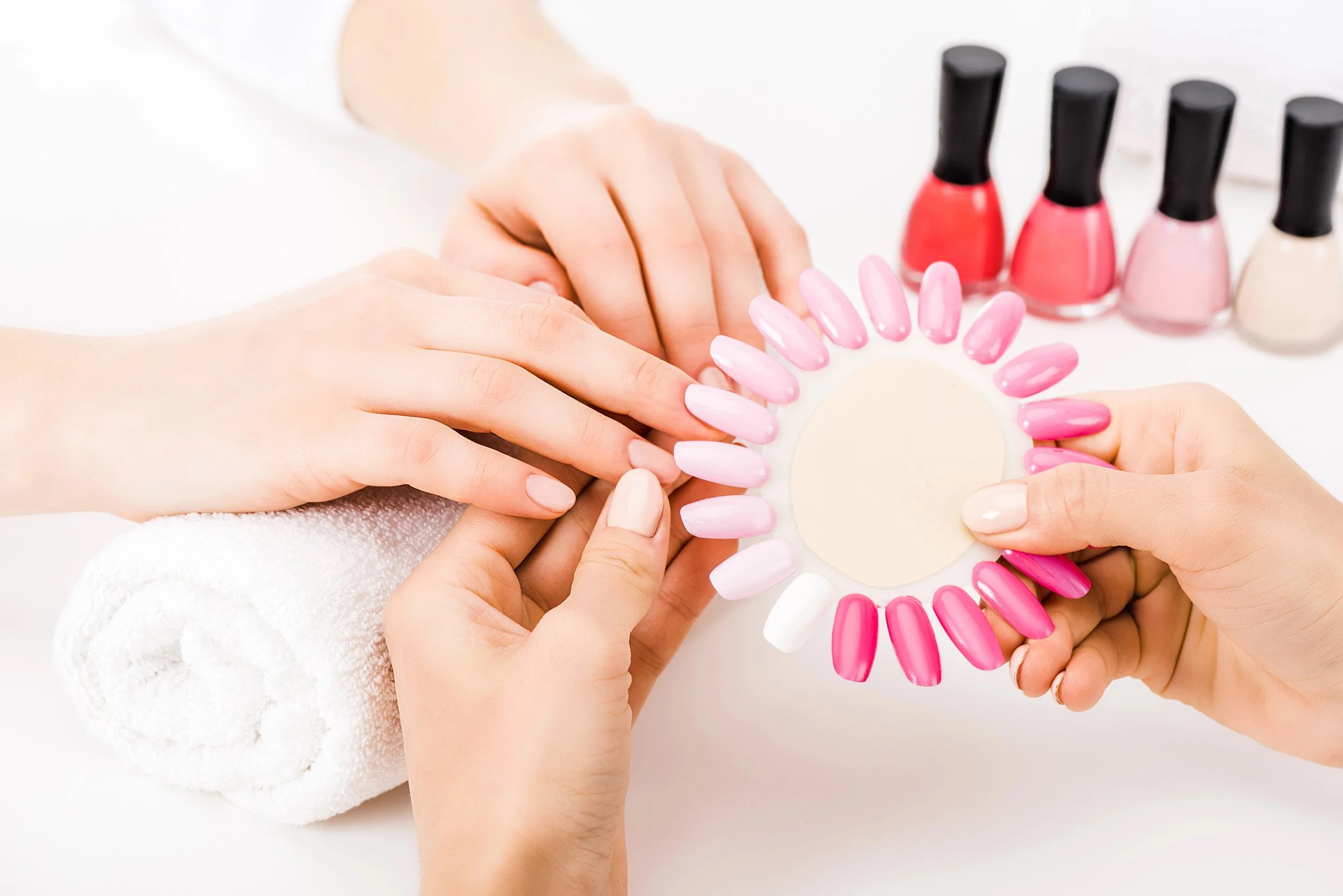 A person getting a manicure, with a hand on a towel and pink and white nail polish samples in a circular holder, and several bottles of nail polish in various shades of pink, red, and white in the background.