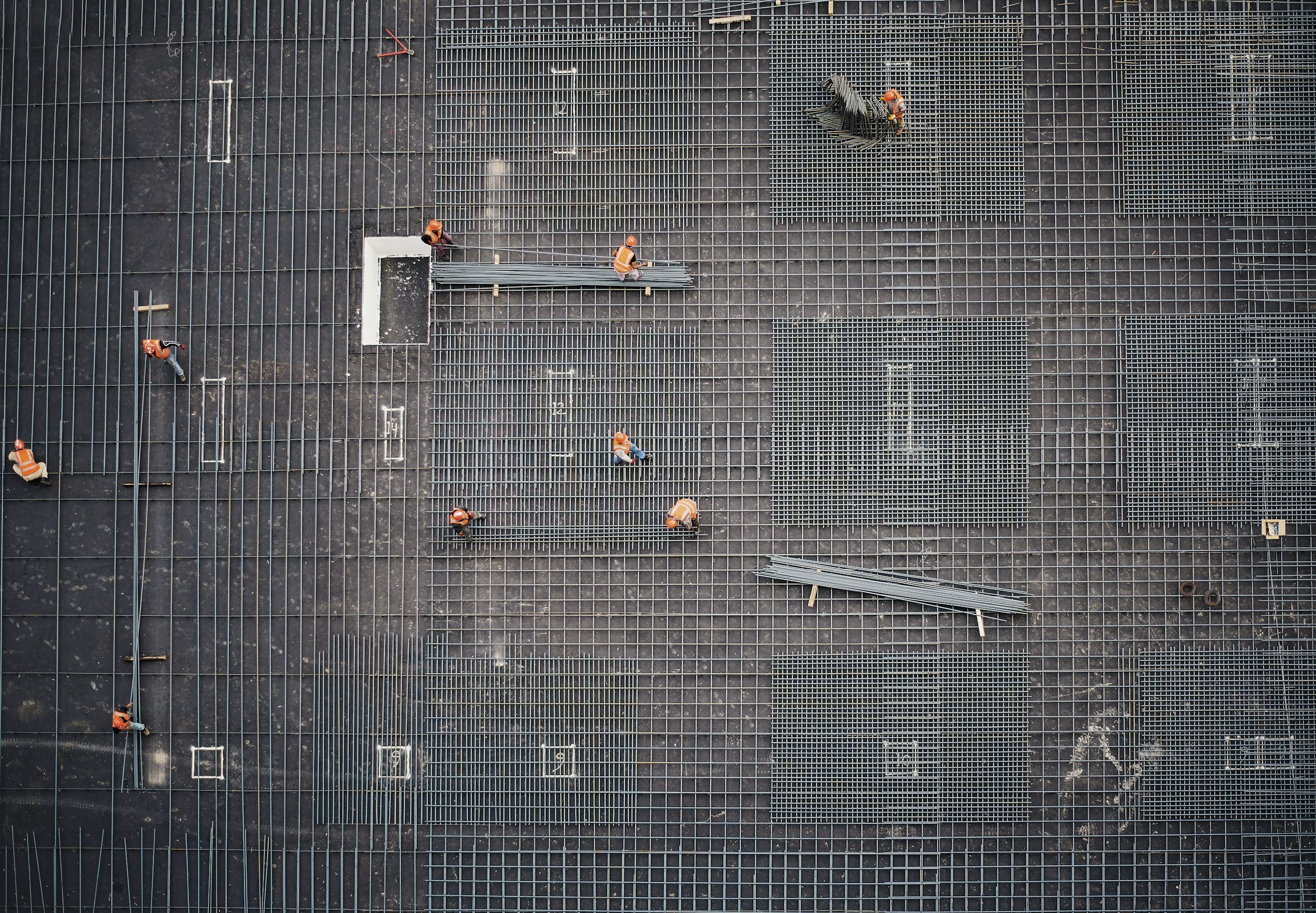 Construction workers in orange safety vests working on building framework with steel grids and metal bars, viewed from above.