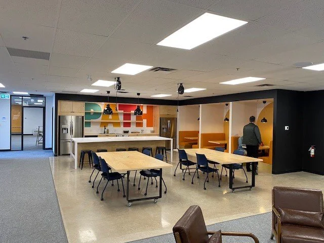 Modern office kitchen and lounge area with tables, chairs, and a person working in a booth.