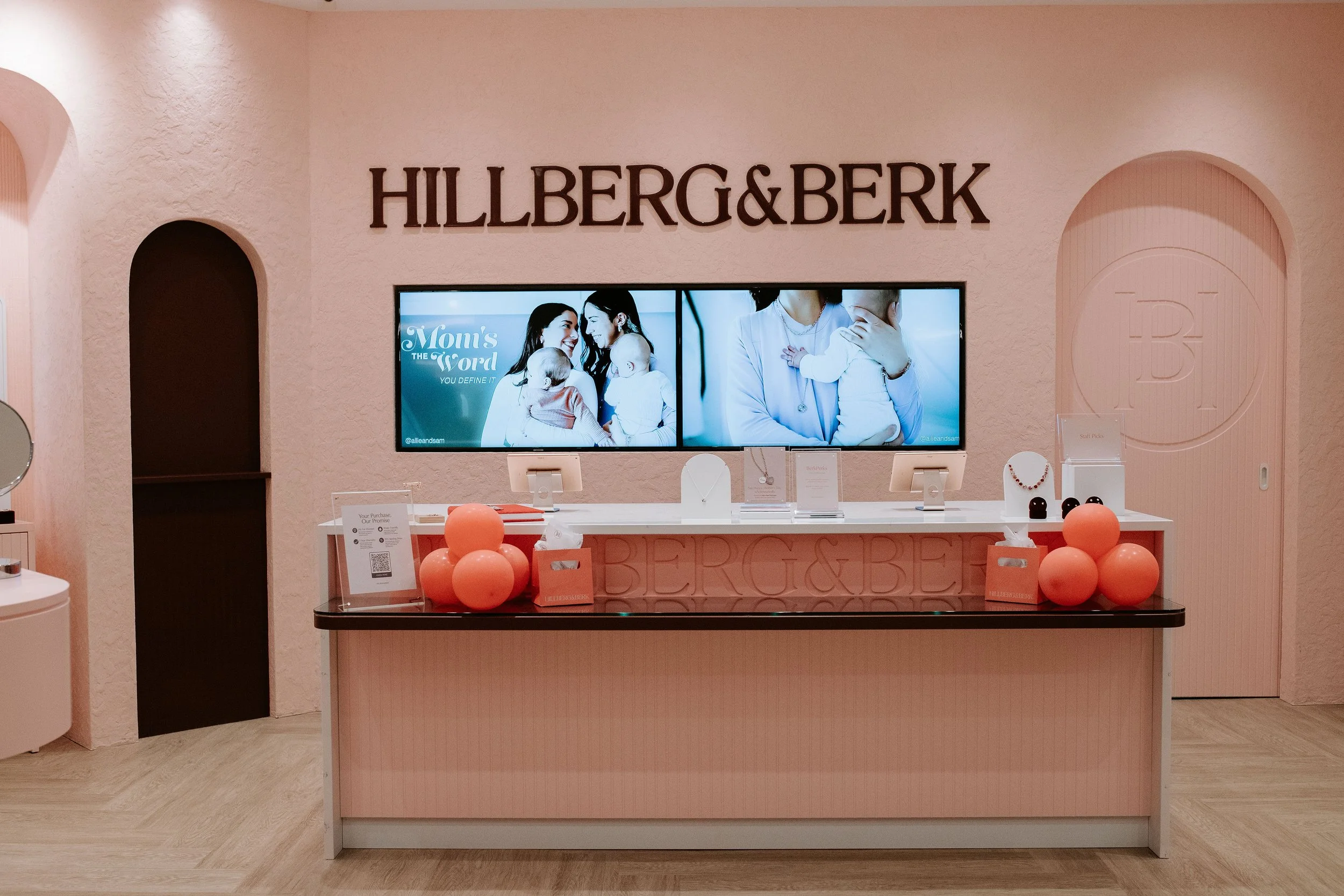 A pink jewelry store boutique with 'HILLBERG & BERK' sign on the wall, a pink checkout counter with balloons, and digital screens showing women with children.
