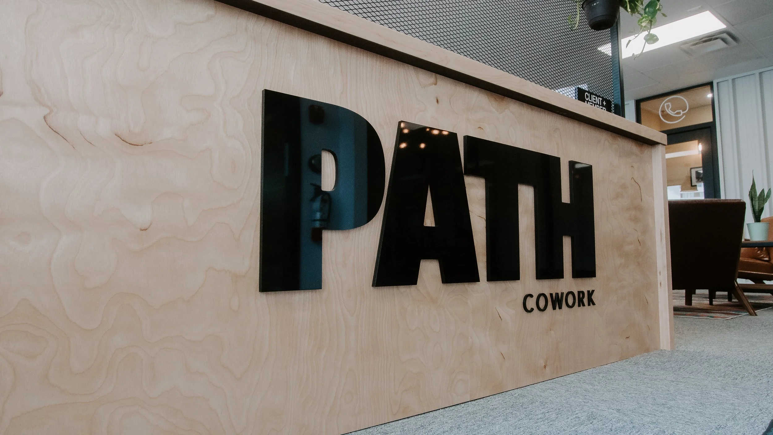 Close-up of a reception desk with a wooden surface and black signage reading 'PATI COWORK' in an office lobby with seating area and plants in the background.