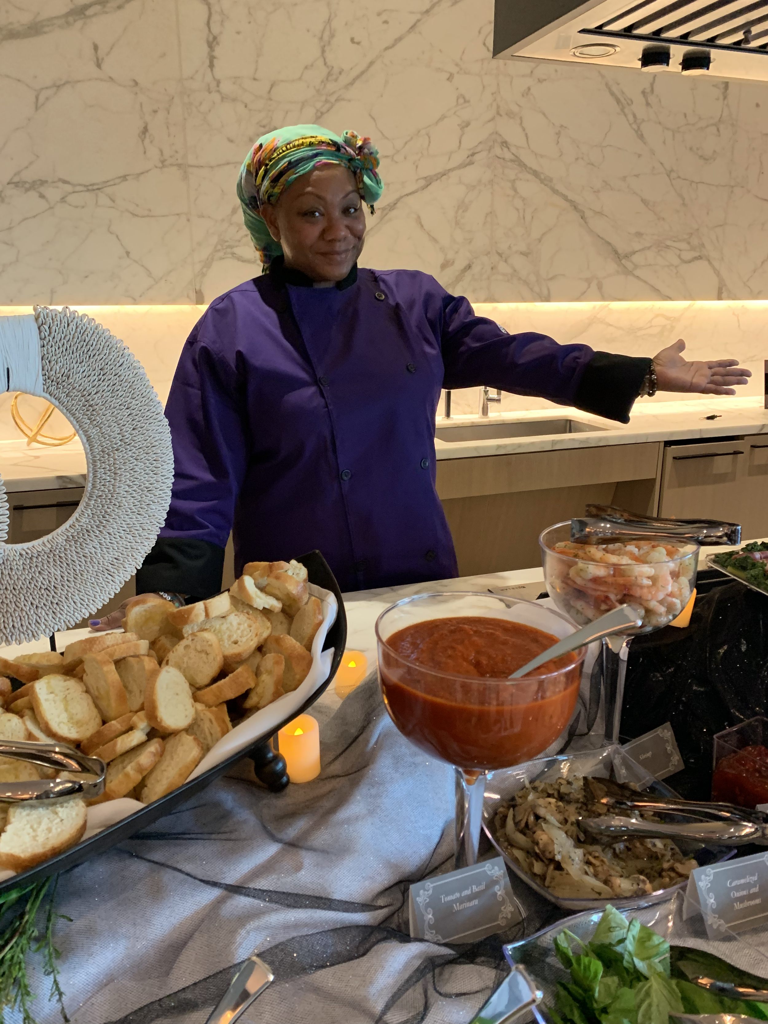 A woman in a purple chef's coat and colorful headscarf stands behind a table of food in a modern kitchen. She is smiling and gesturing towards the spread, which includes bread, shrimp, tomato sauce, and other dishes.