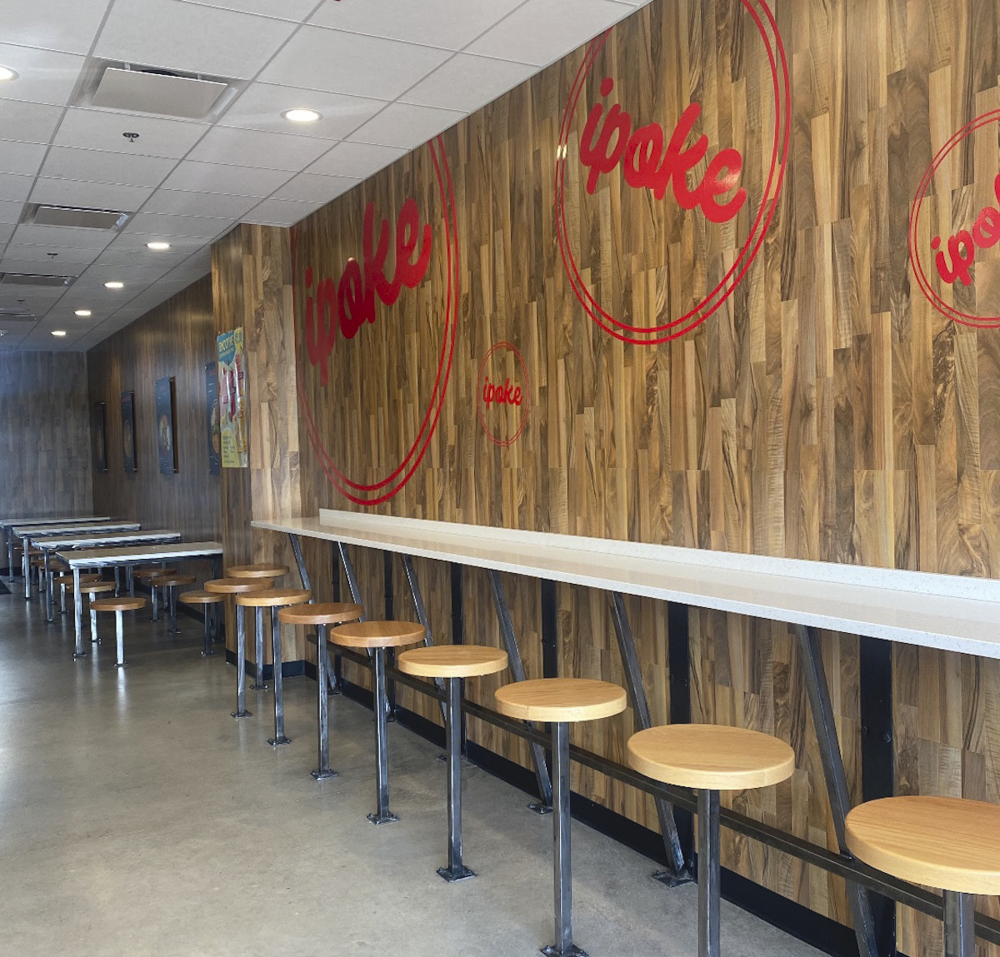 Empty cafe or restaurant seating area with a row of wooden stools attached to a long white counter along a wood-paneled wall featuring red circular text that reads 'ipoke'.