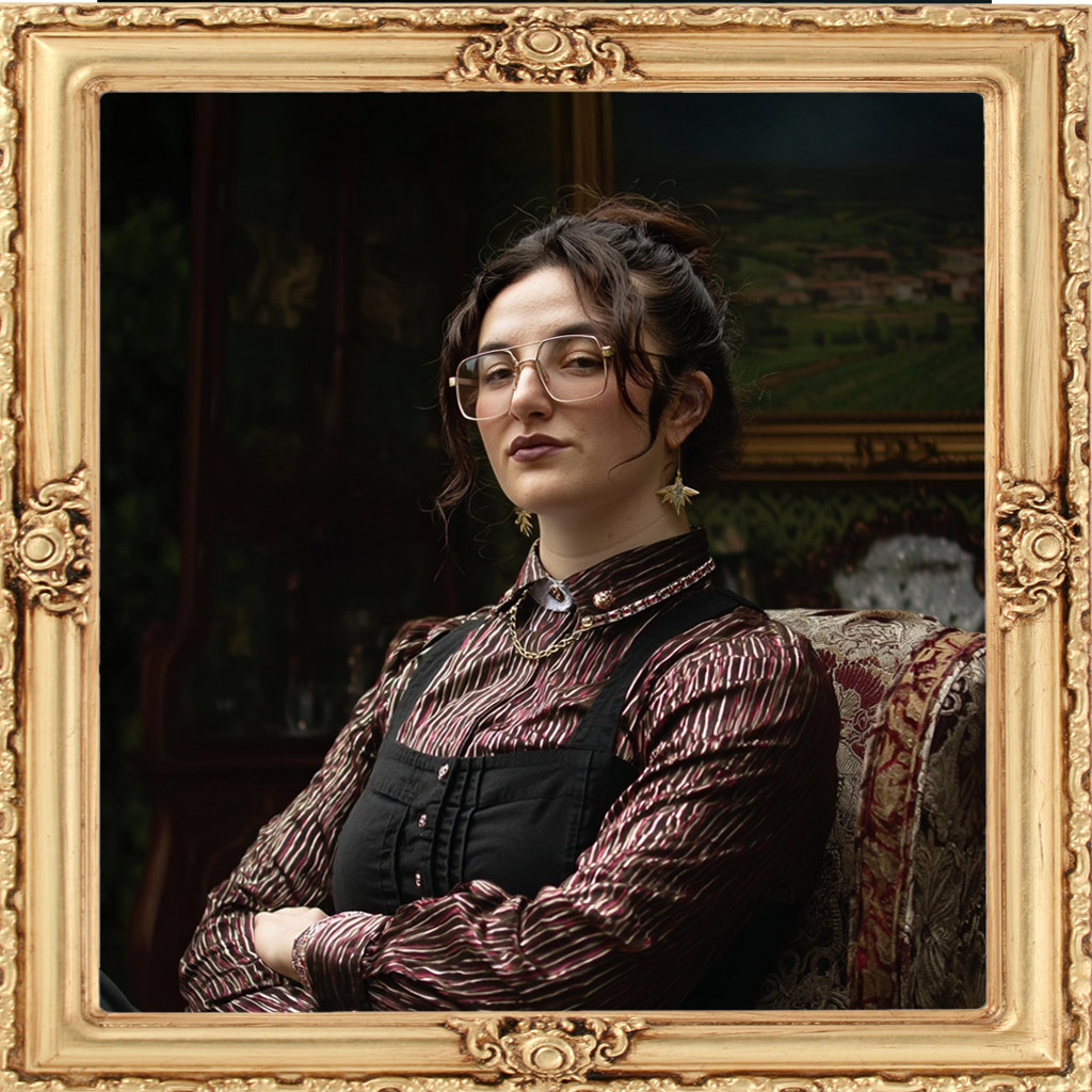 A woman with dark hair, glasses, and earrings sitting in an ornate chair, cross-armed, wearing a patterned blouse and black overalls, in front of a dark background with a framed landscape painting.