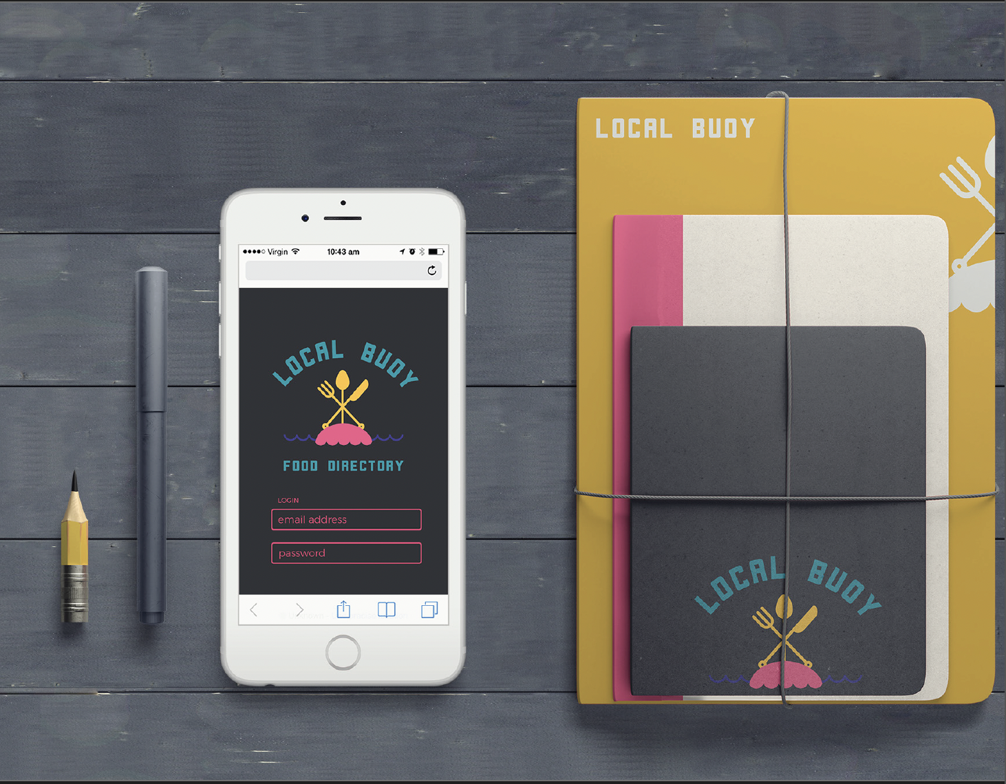 A smartphone displaying the 'Local Buoy' food directory app next to a yellow pen, a gray pen, and three notebooks in pink, gray, and black on a dark wood surface.