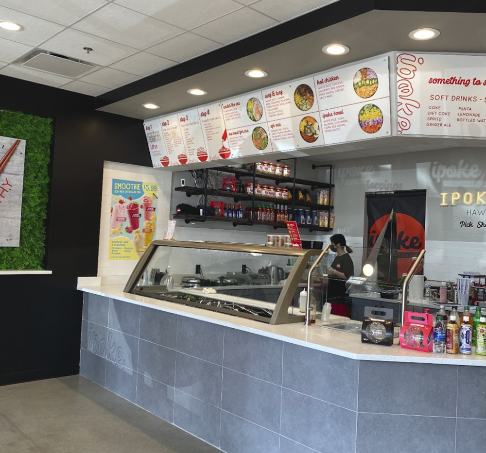 Counter at a fast food restaurant with a glass display case, menu above, and shelves with condiments and supplies behind the counter.