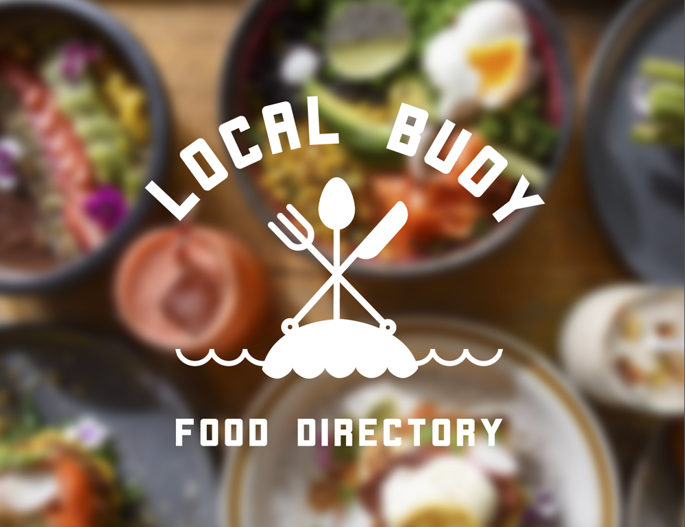 Overhead view of various bowls of colorful salads and food dishes on a wooden table, with a logo in the center that reads 'Local Food Buddy Food Directory'.