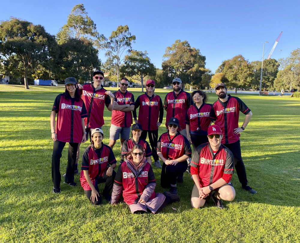 A group of eleven people wearing matching red and black sports jerseys with 'BUNTFORCE' on them, posing outdoors on a grassy field with trees and a clear blue sky in the background.