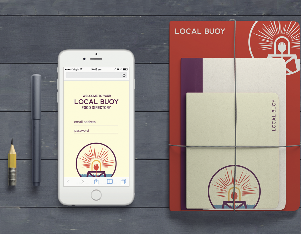 A smartphone displaying a food directory app, two pens, and a set of four notebooks with a 'Local Buoy' logo, all on a dark wooden surface.