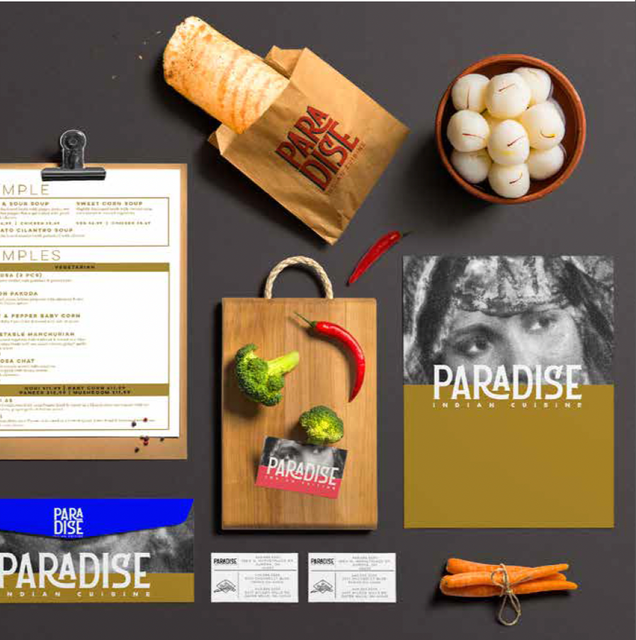 Flat lay of Indian cuisine items including a paper bag with the words "Dare to dish," a bowl of white onions, a wooden cutting board with broccoli and red chili peppers, a menu on a clipboard, and promotional materials for Paradise Indian Cuisine on a dark surface.