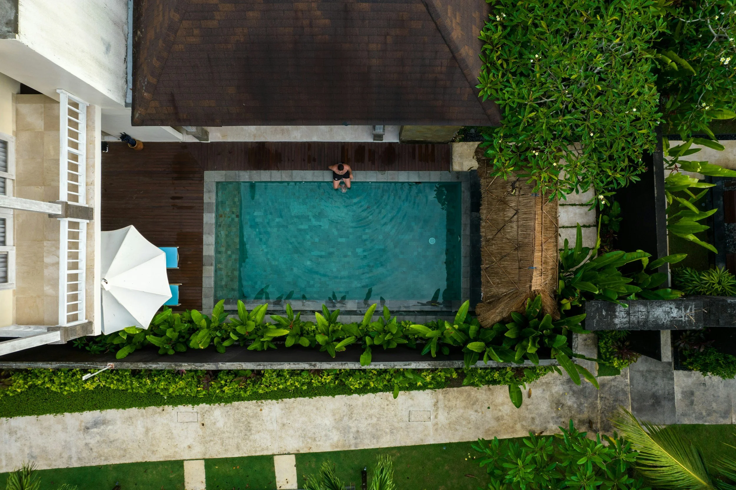 Aerial view of a small swimming pool adjacent to a wooden deck, with a person standing at the edge of the pool, surrounded by lush greenery and outdoor furniture.