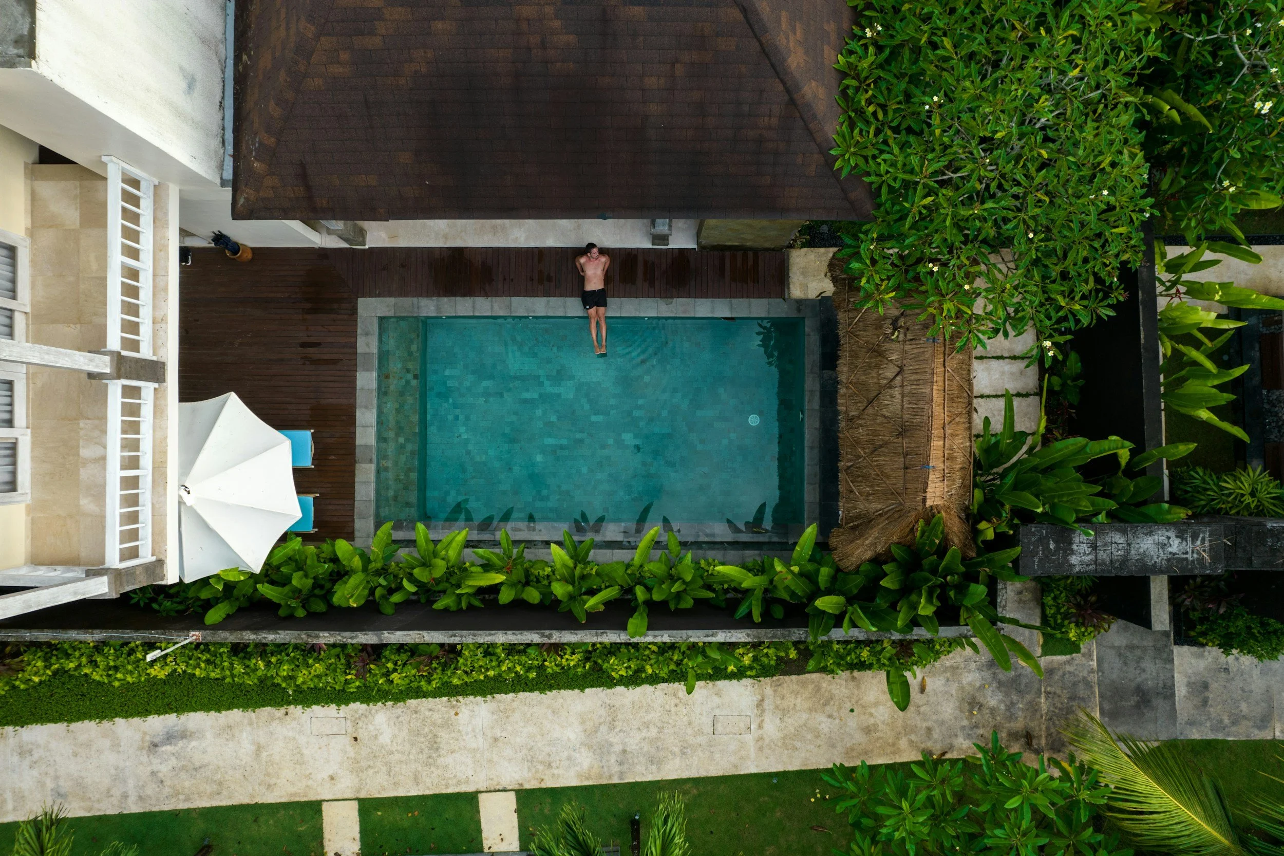 An overhead view of a person lying on a swimming pool's edge with arms behind their head. The pool is surrounded by lush greenery, wooden decking, and white umbrellas.