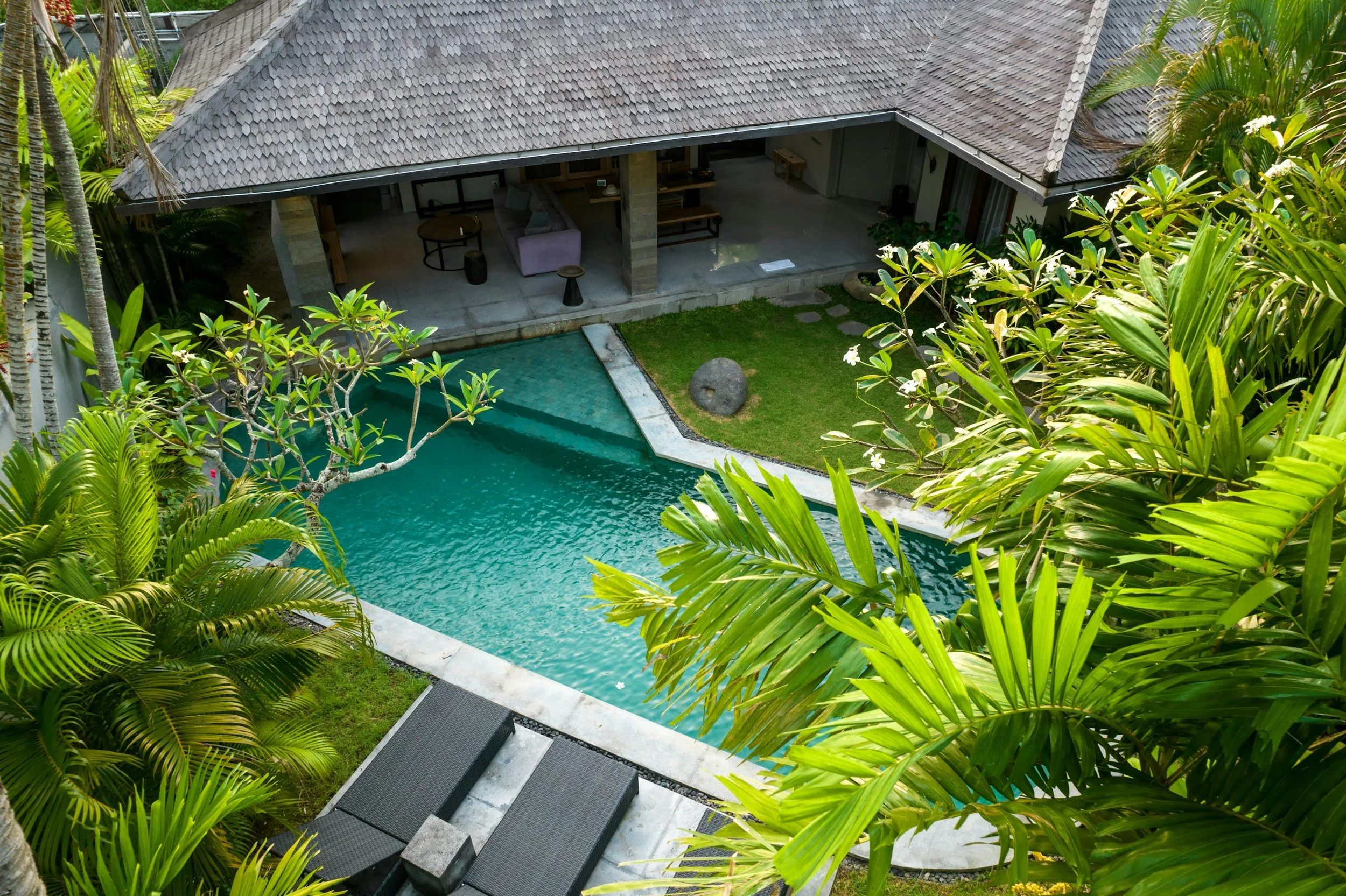 Aerial view of a swimming pool surrounded by tropical plants and trees, with a covered patio area featuring lounge chairs and outdoor furniture.