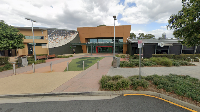 The entrance to Upper Coomera Centre with a pathway leading to a modern building under a cloudy sky.