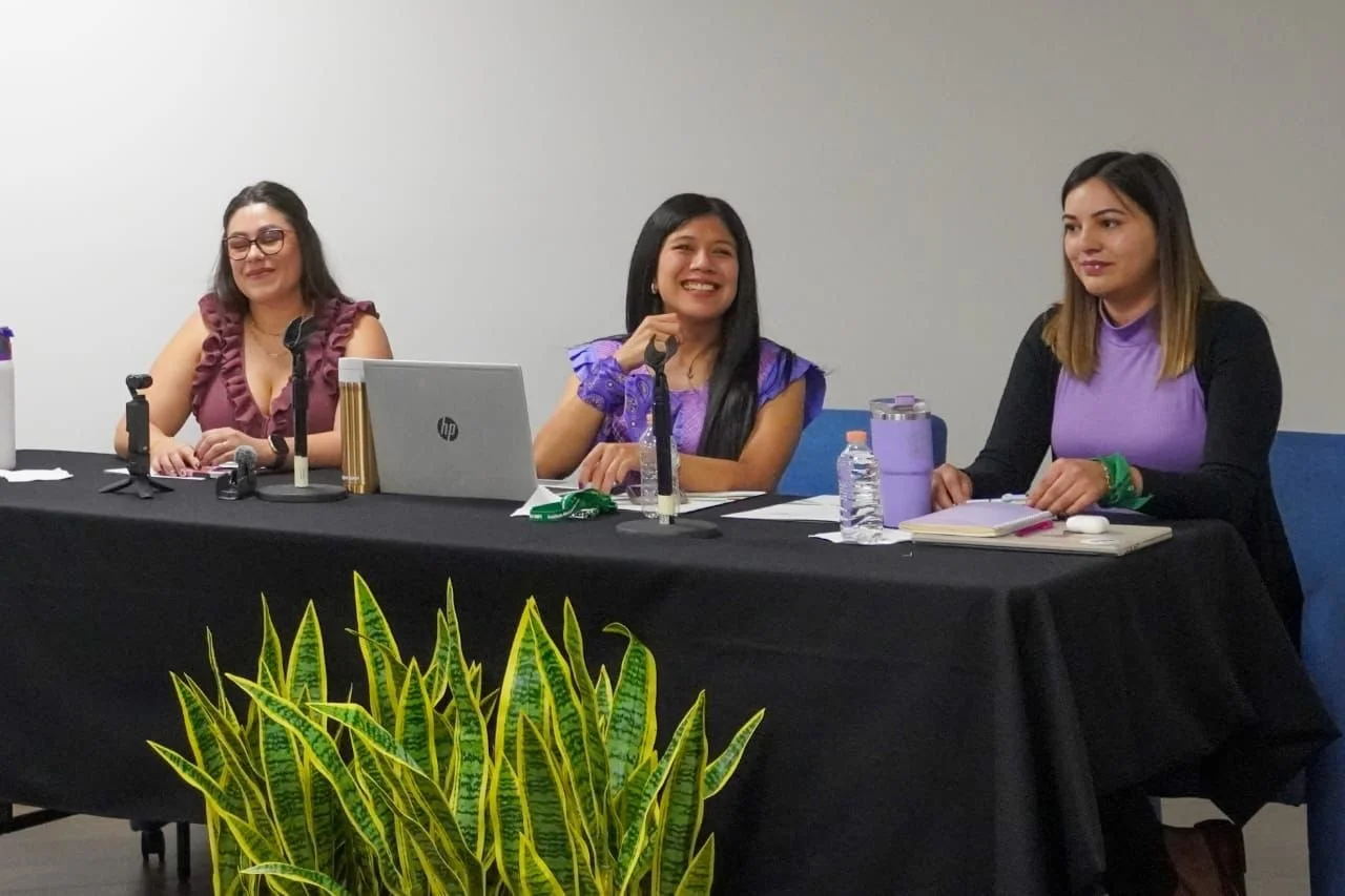 Tres mujeres sentadas en una mesa en una conferencia, con micrófonos, laptops y botellas de agua.