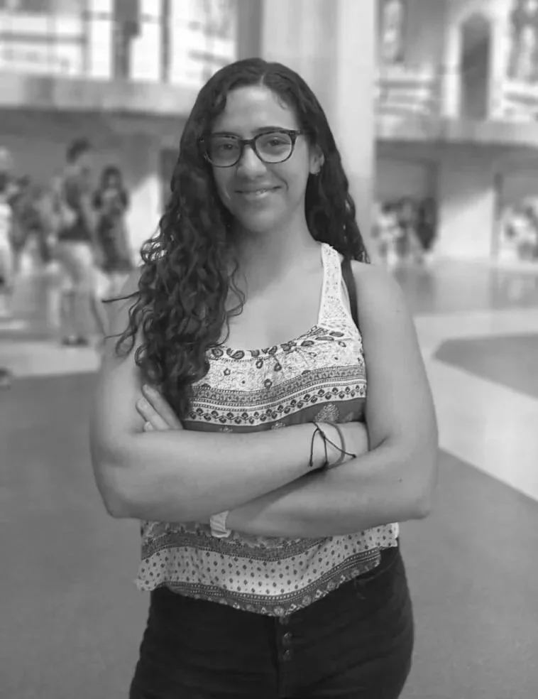 Joven mujer de cabello rizado y gafas, sonriendo y con brazos cruzados, en un espacio público con varias personas de fondo.