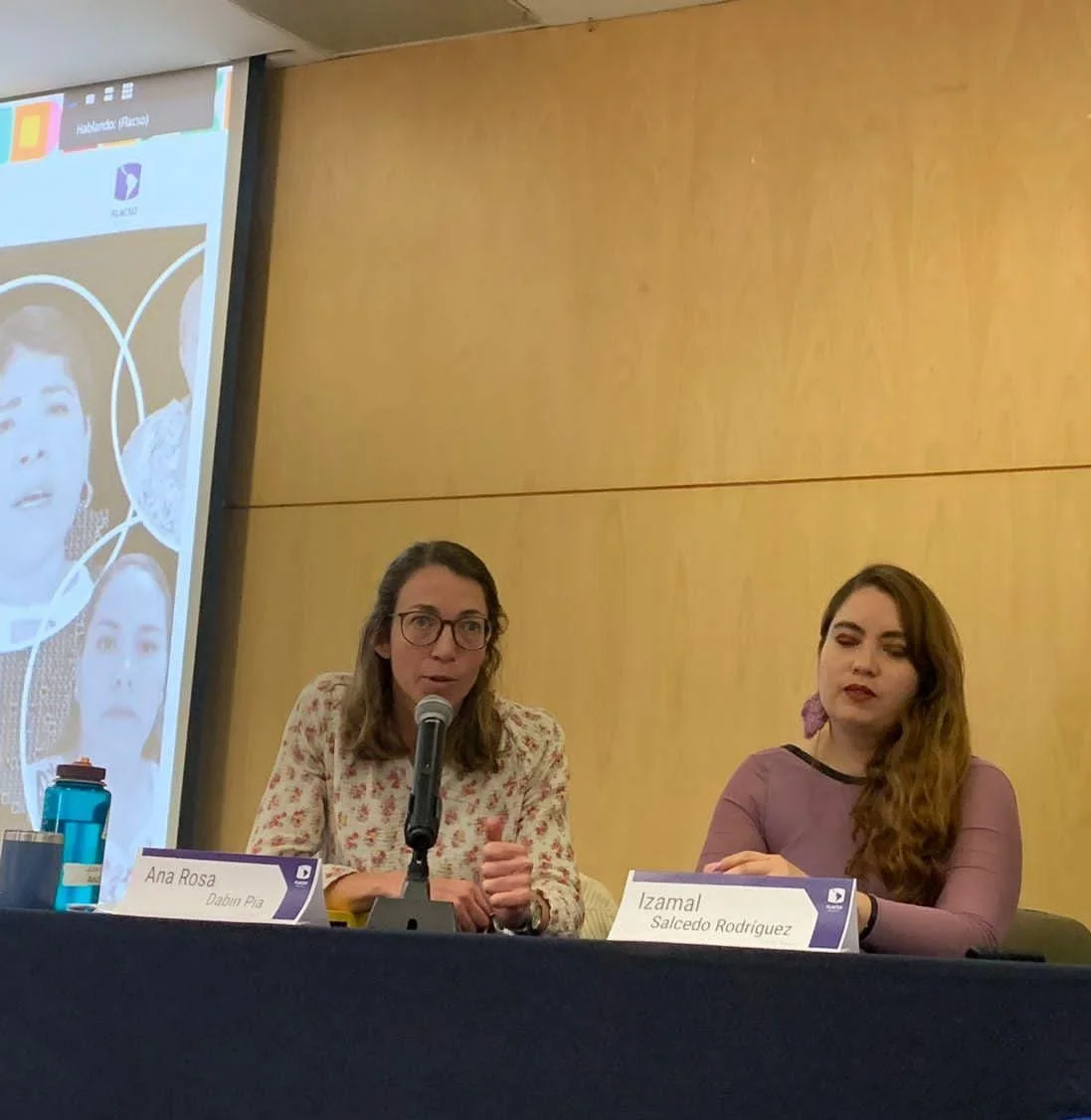 Dos mujeres sentadas en una mesa en una conferencia. La mujer a la izquierda habla, lleva gafas y una blusa con estampado floral. La mujer a la derecha tiene cabello rizado, lleva una blusa lila y está escuchando. Hay un nombre y un cartel en la mesa que dicen Ana Rosa Dabiín P3 y Izamal Salcedo Rodríguez. En el fondo, una pantalla con imágenes y texto.