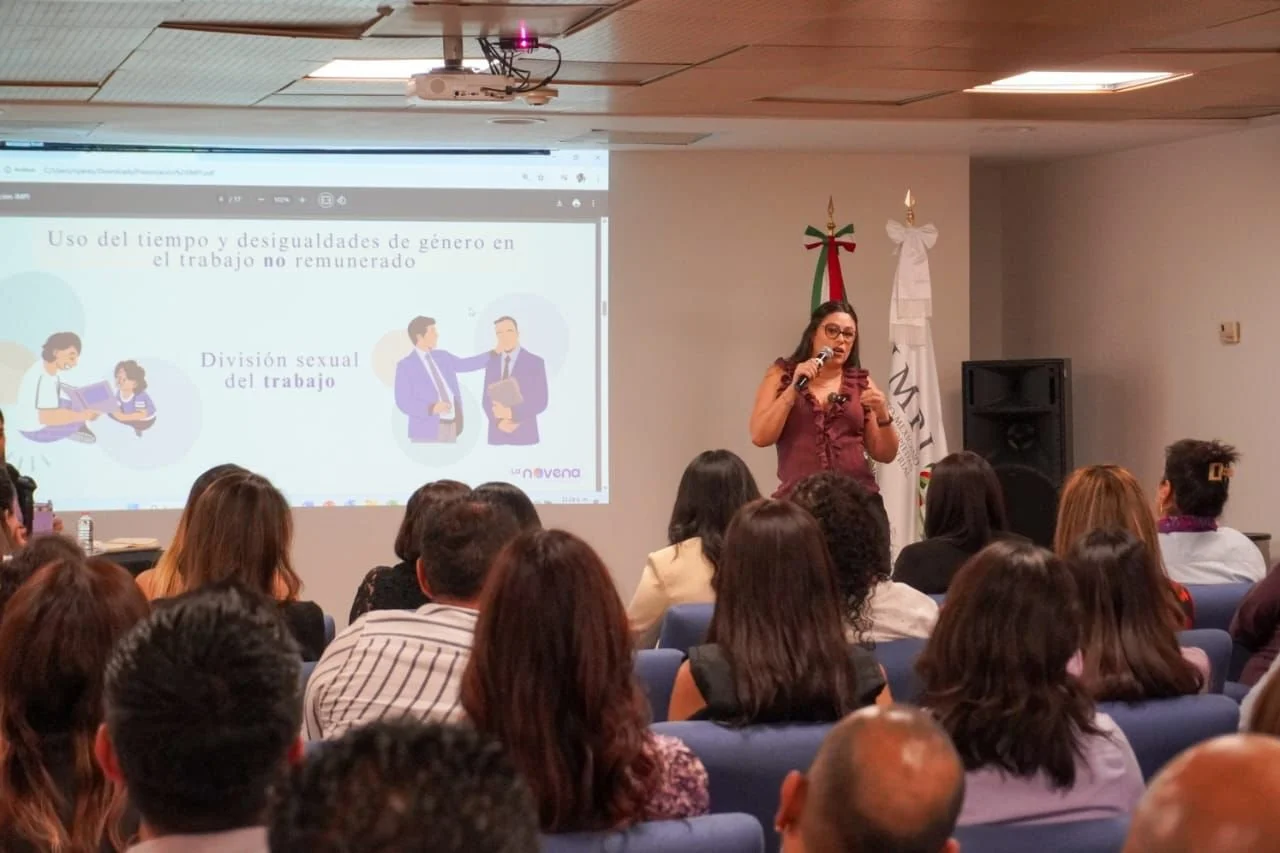Mujer dando una charla en una conferencia con una presentación en una pantalla que habla sobre desigualdades de género y división sexual del trabajo, con un público sentado en sillas azules.