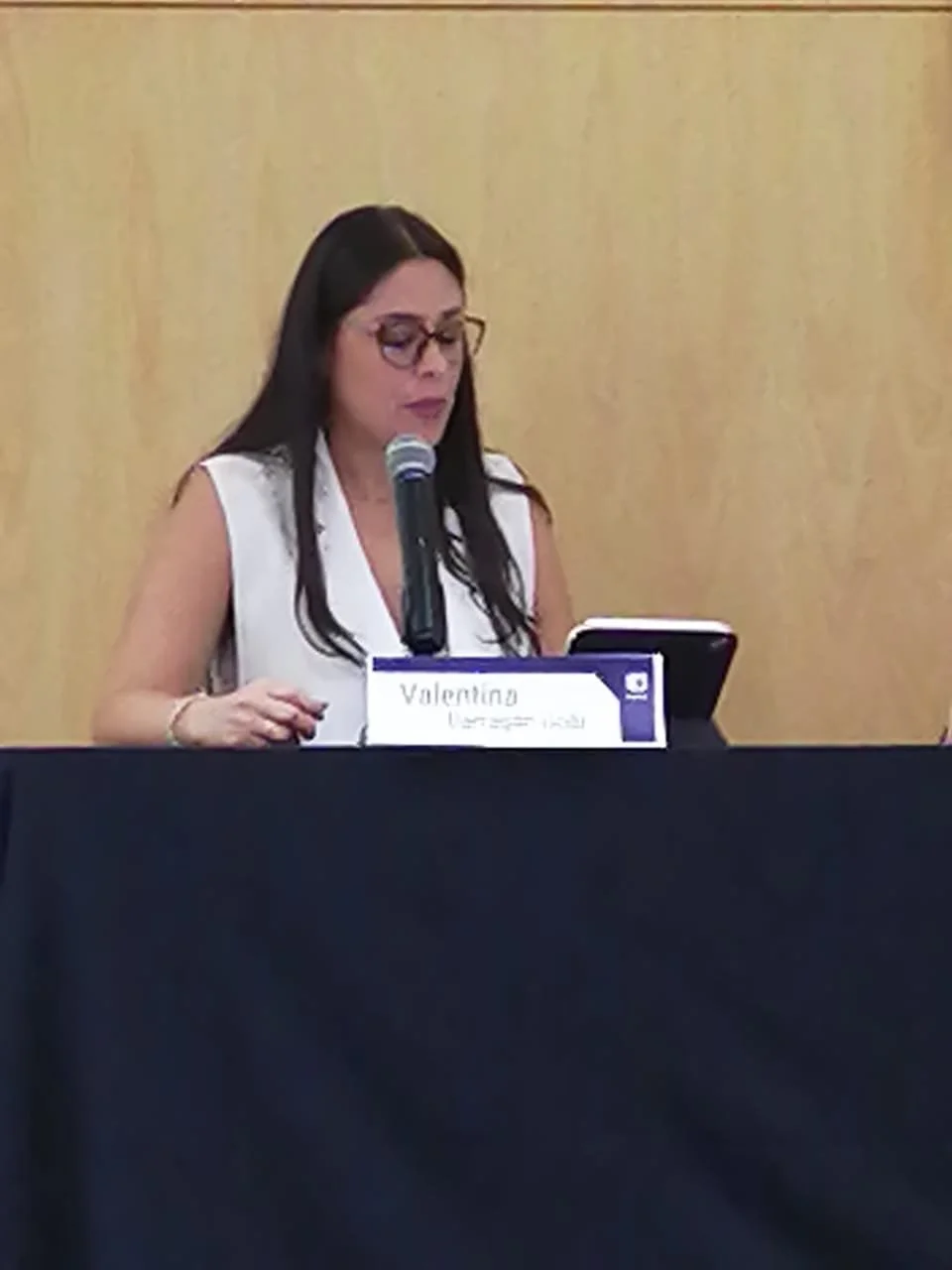 Mujer con gafas, de cabello largo oscuro, sentada en una mesa con un micrófono y una tableta, en un entorno de conferencia o reunión.