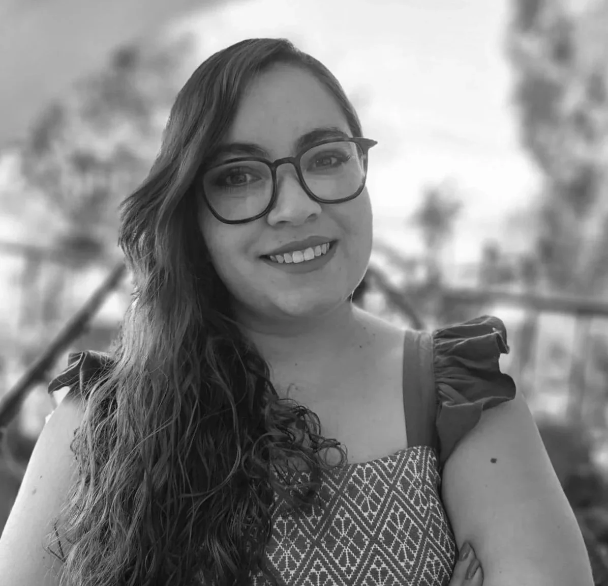 Retrato en blanco y negro de una mujer sonriendo, con gafas grandes, cabello rizado y vestido con patrón geométrico, en un entorno al aire libre con árboles de fondo.