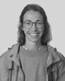 Mujer con gafas sonriendo, lleva una chaqueta y un suéter, fondo blanco y simple.