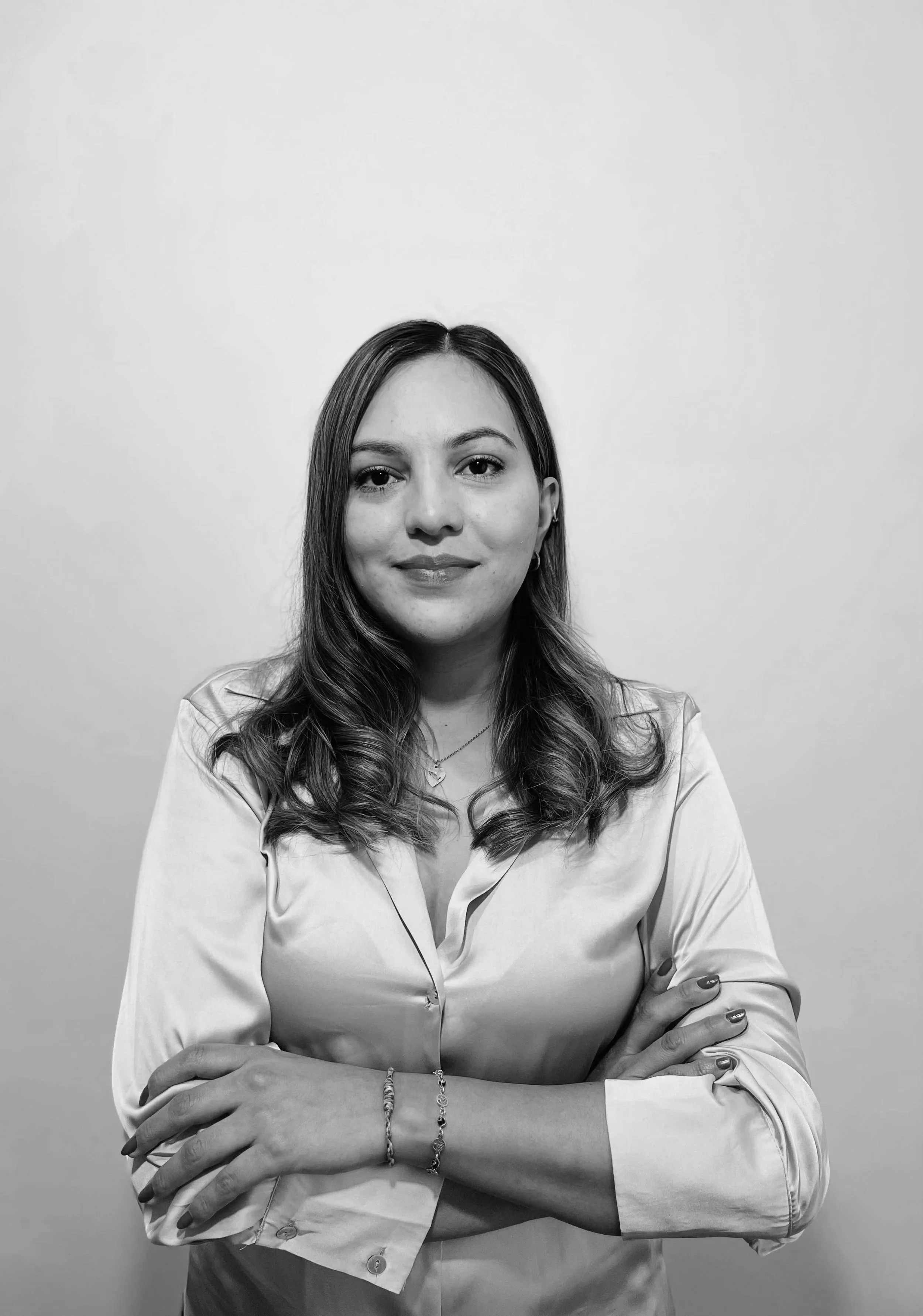 Retrato de una mujer con cabello castaño claro y rizado, usa blusa de manga larga y accesorios, mira a la cámara con expresión neutral, fondo blanco.