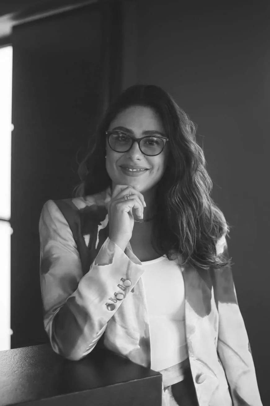 Retrato en blanco y negro de una mujer con gafas, cabello rizado, sonriendo y posando con la mano en el mentón, con fondo oscuro.