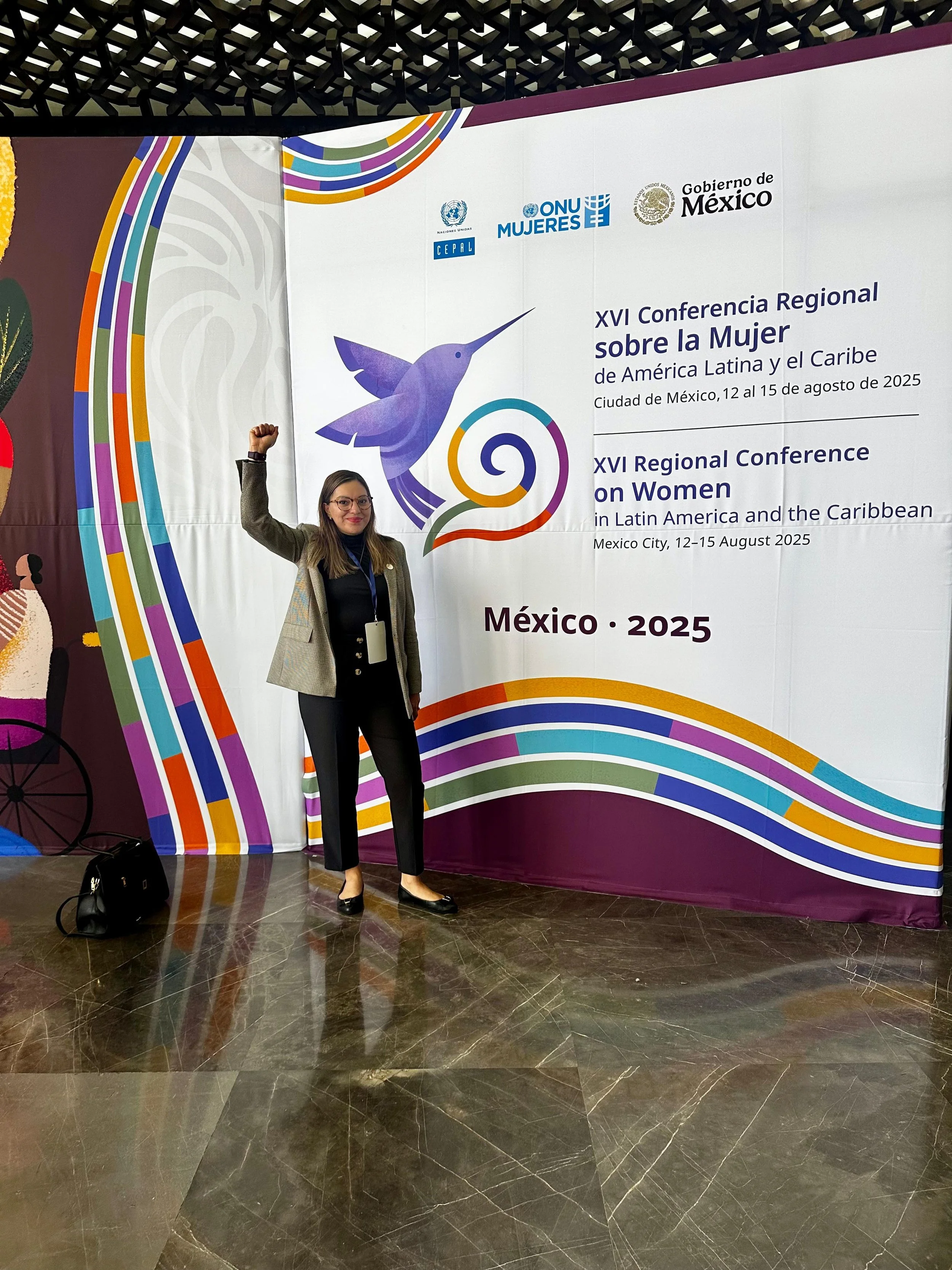 Mujer de pie frente a un cartel en un evento, levantando el puño en señal de victoria, en una conferencia regional en Ciudad de México en agosto de 2025