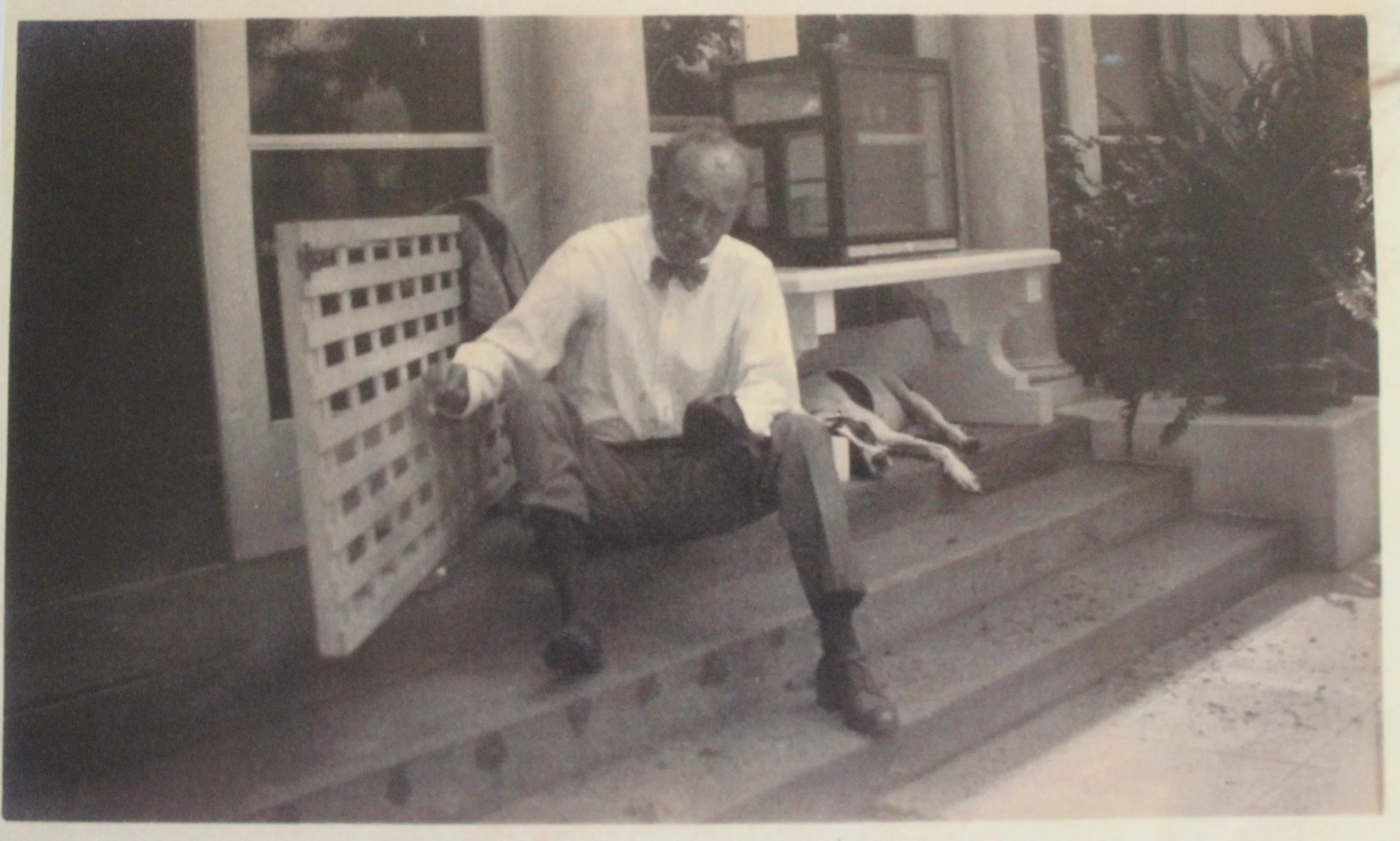 An elderly man wearing glasses and a bowtie sitting on a wooden park bench with a dog lying beside him. The setting appears to be outdoors, with trees or bushes visible in the background.