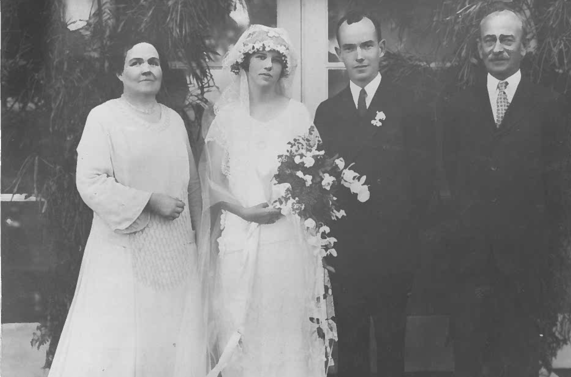 A black and white photo of four people, likely a family, standing outdoors with trees and plants in the background. From left to right, there is a woman with dark hair, an individual dressed in a wedding gown with a floral headpiece, a man in a tuxed