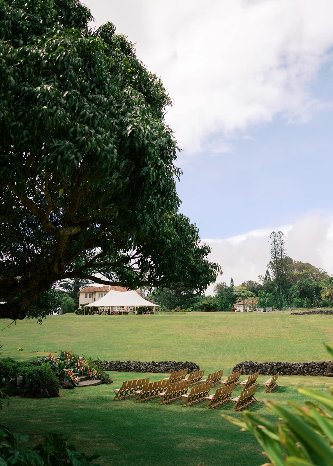 A large green lawn with rows of wooden chairs set up for an outdoor event, trees and a white canopy in the background, and a house and other buildings visible beyond the lawn under a partly cloudy sky.