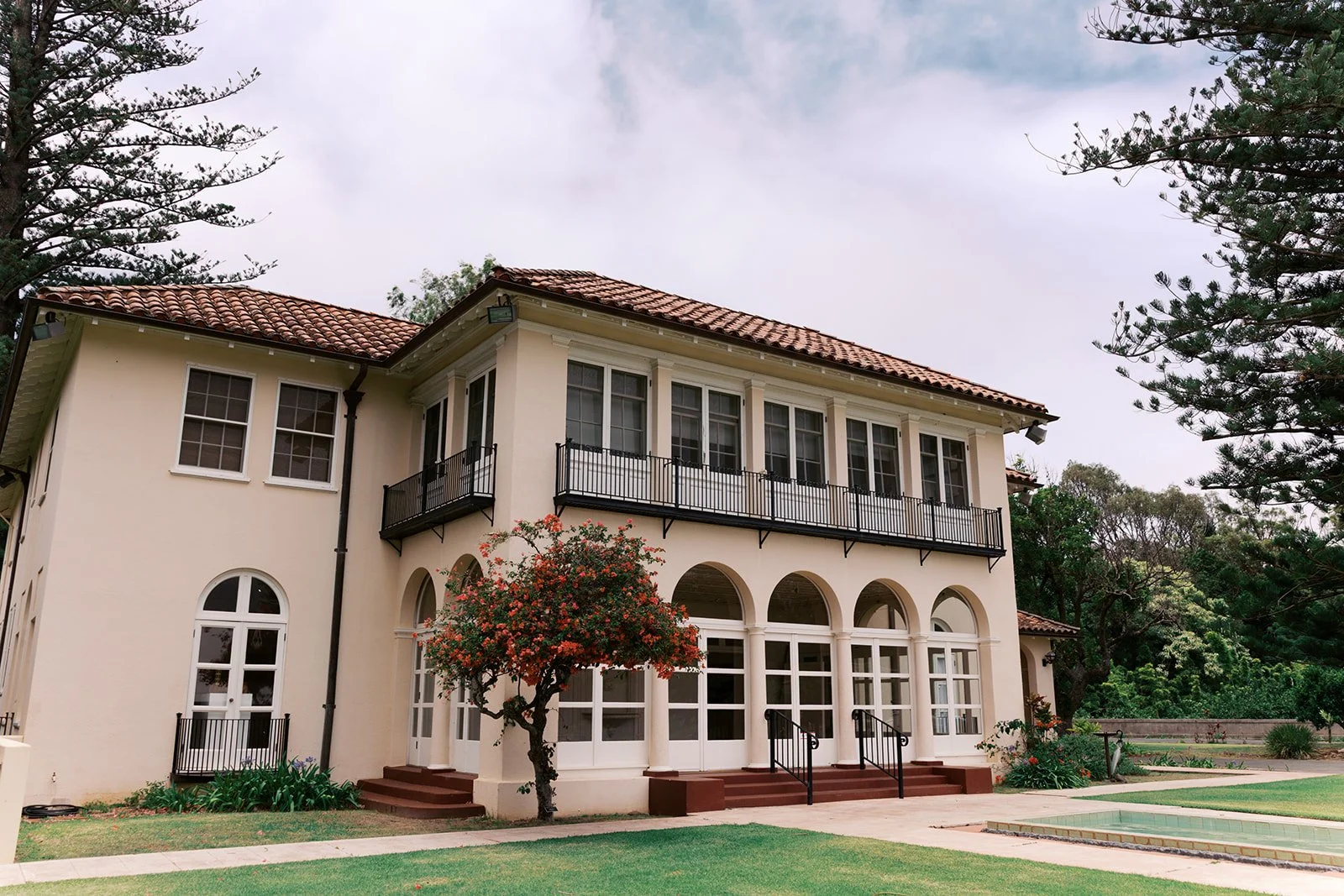 A two-story house with a beige exterior, red tiled roof, and multiple windows. The house has a balcony on the upper level with black railings and an arched porch on the lower level. There is a small tree with pink flowers in front, and the yard has g