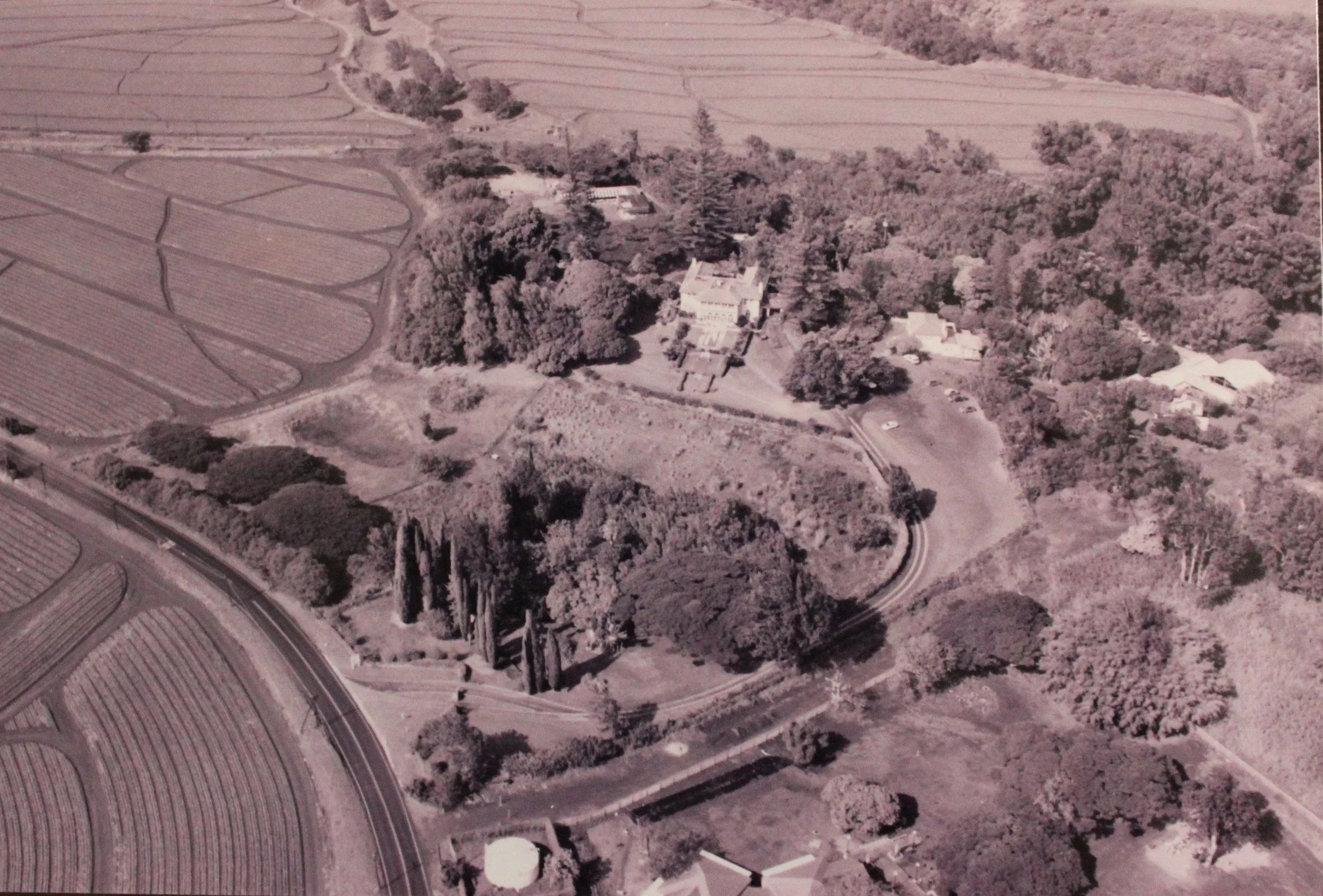 An aerial view of farmland with fields, trees, and roads surrounding a large estate or mansion. There are also some smaller buildings and a parking area visible.
