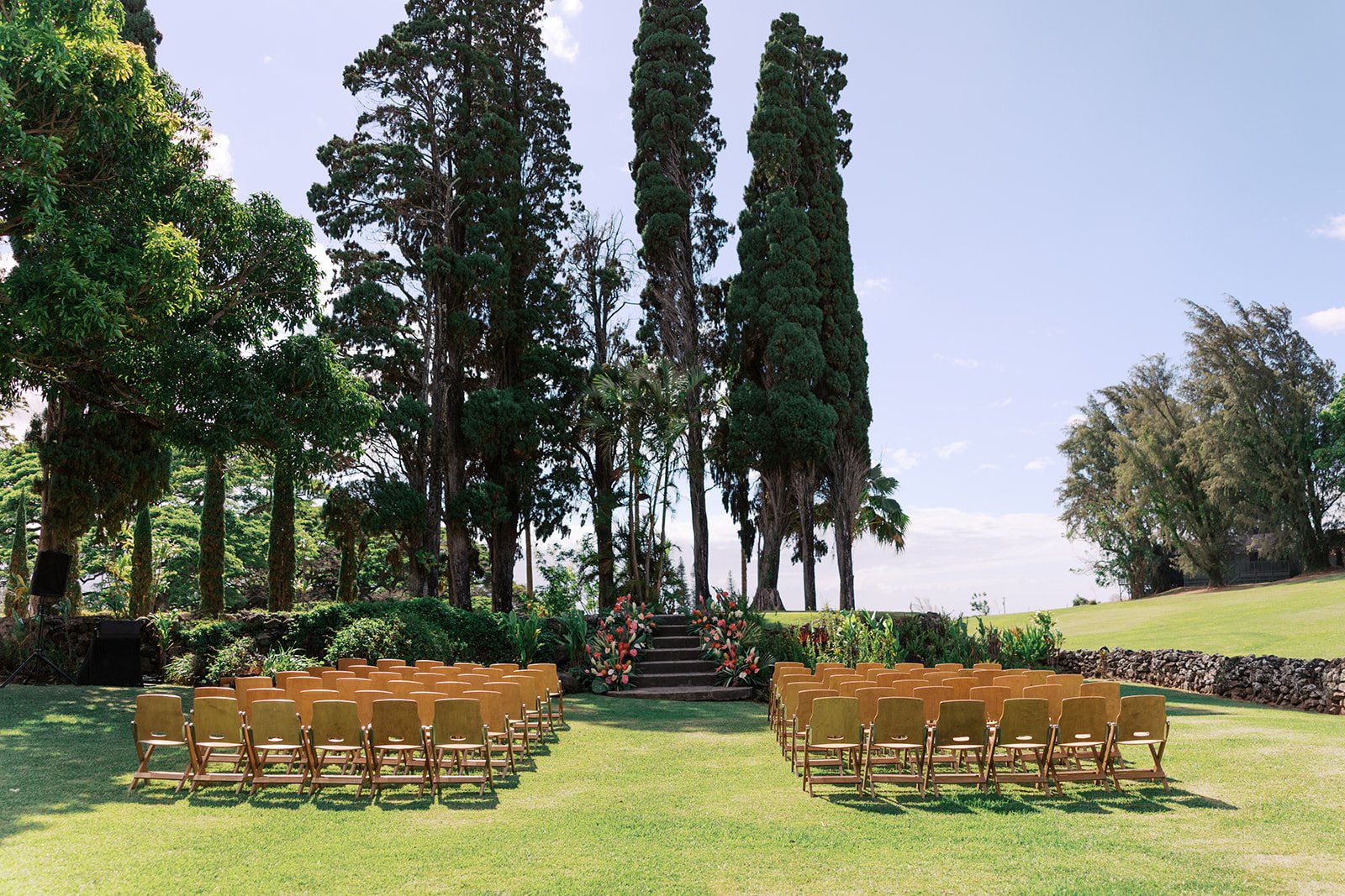 An outdoor wedding setup with rows of chairs facing a floral altar on steps, surrounded by trees and lush greenery under a bright blue sky.