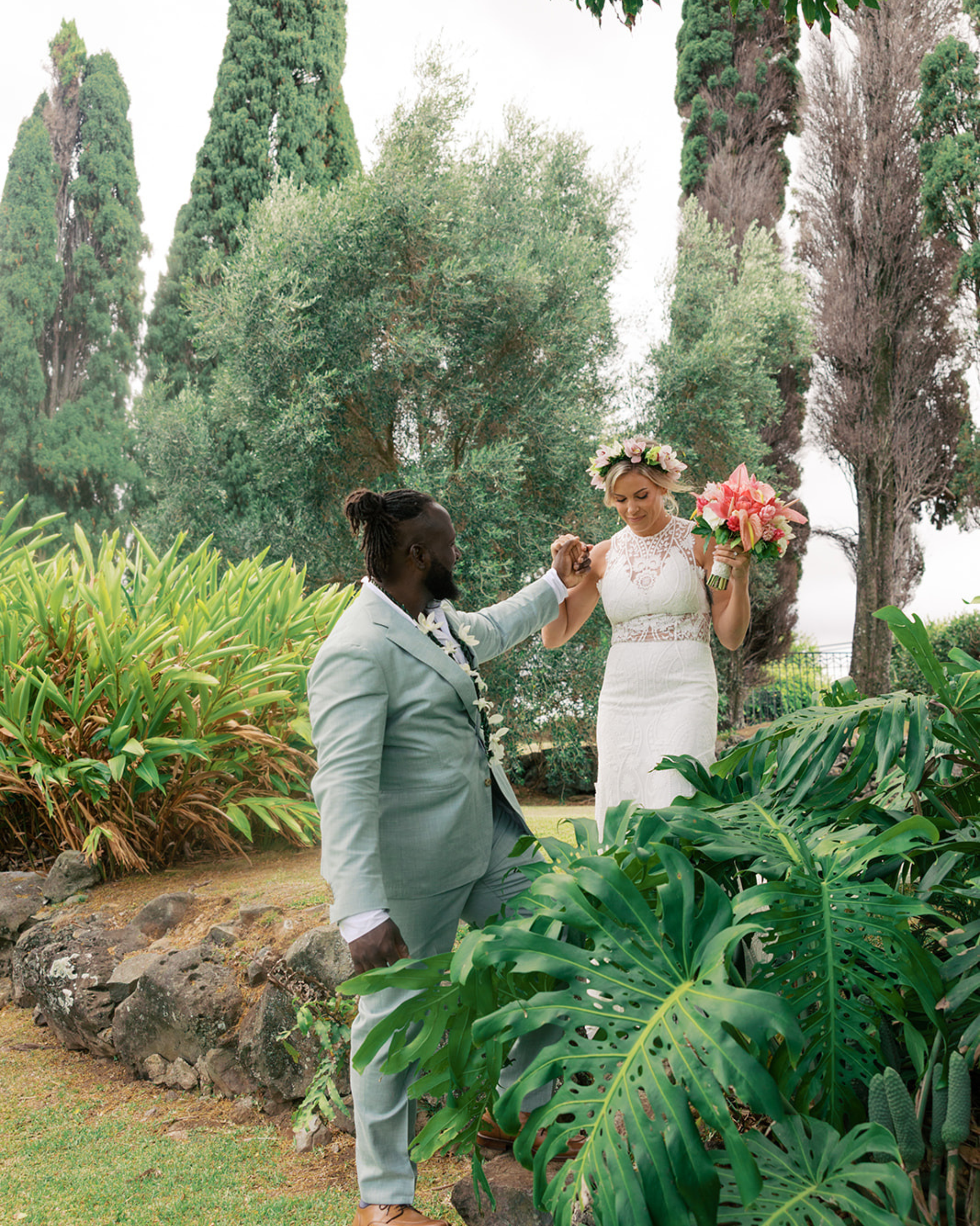 A man kneeling on the grass and holding the hand of a woman standing beside him, with a lush garden and tall trees in the background. The woman is holding a colorful bouquet of flowers and wearing a floral crown.