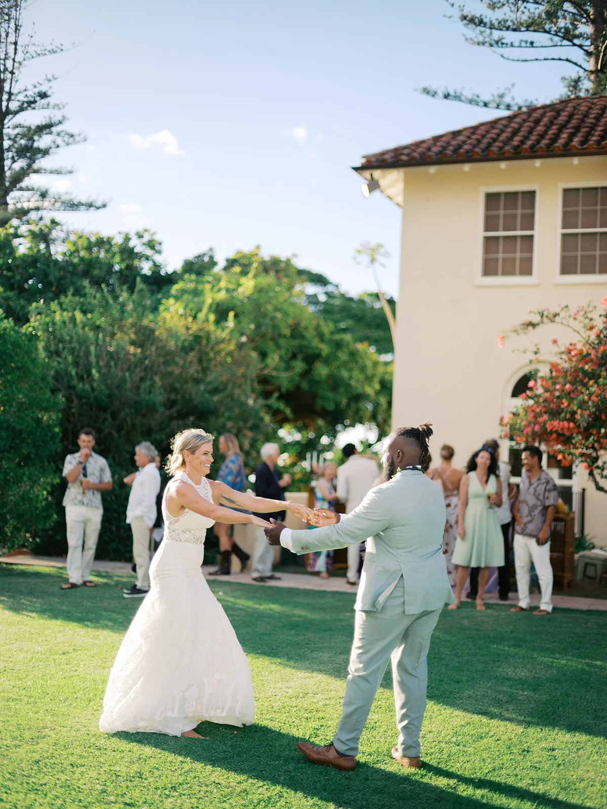 A bride and groom dance together on a lawn at an outdoor wedding reception, with guests in the background near a yellow house and lush greenery.