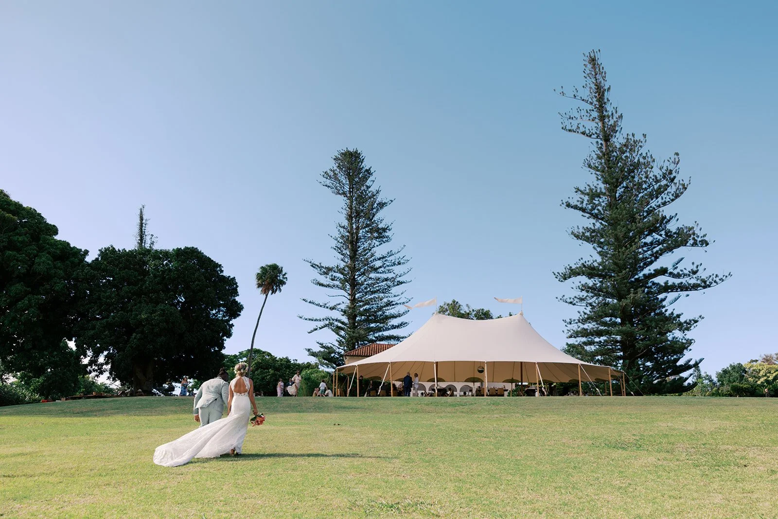 A bride and groom walking on a grassy wedding venue with a large white tent, tall trees, and a clear blue sky in the background.