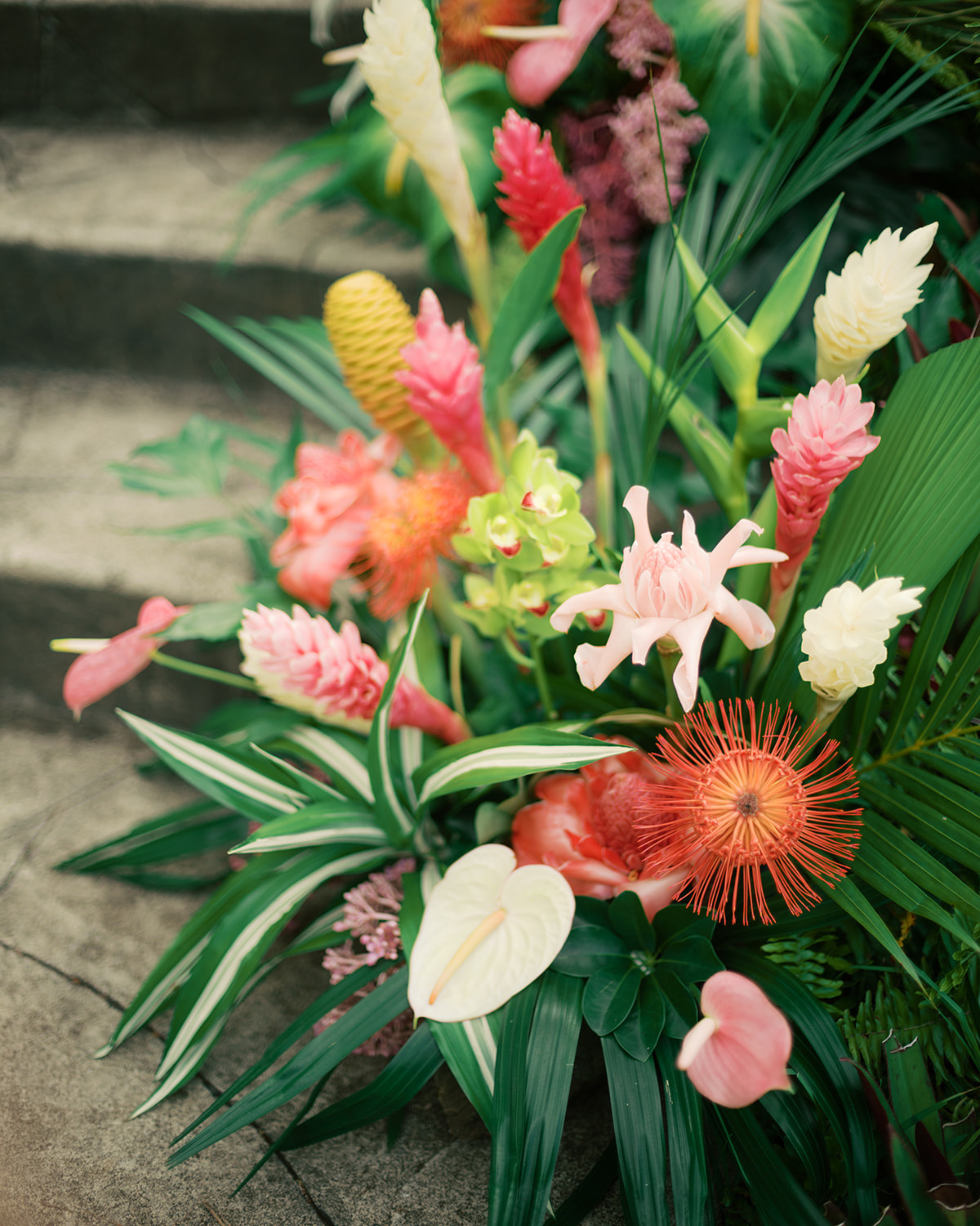 Colorful tropical flower arrangement with pink, yellow, white, and red flowers and green leafy foliage on a concrete surface.