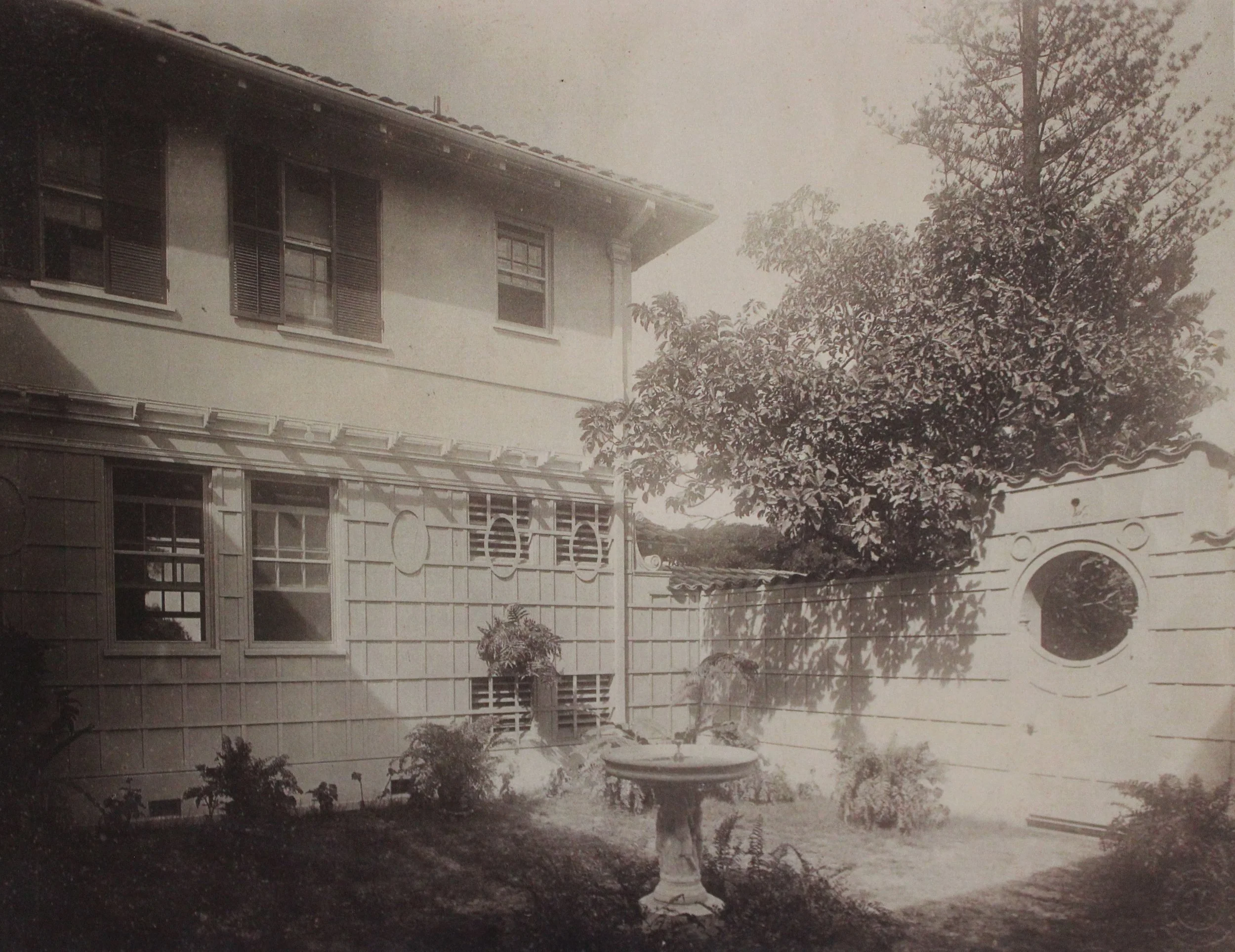 A sepia-toned photograph of a two-story house with multiple windows, surrounded by trees and a garden, and a curved gate in the foreground.