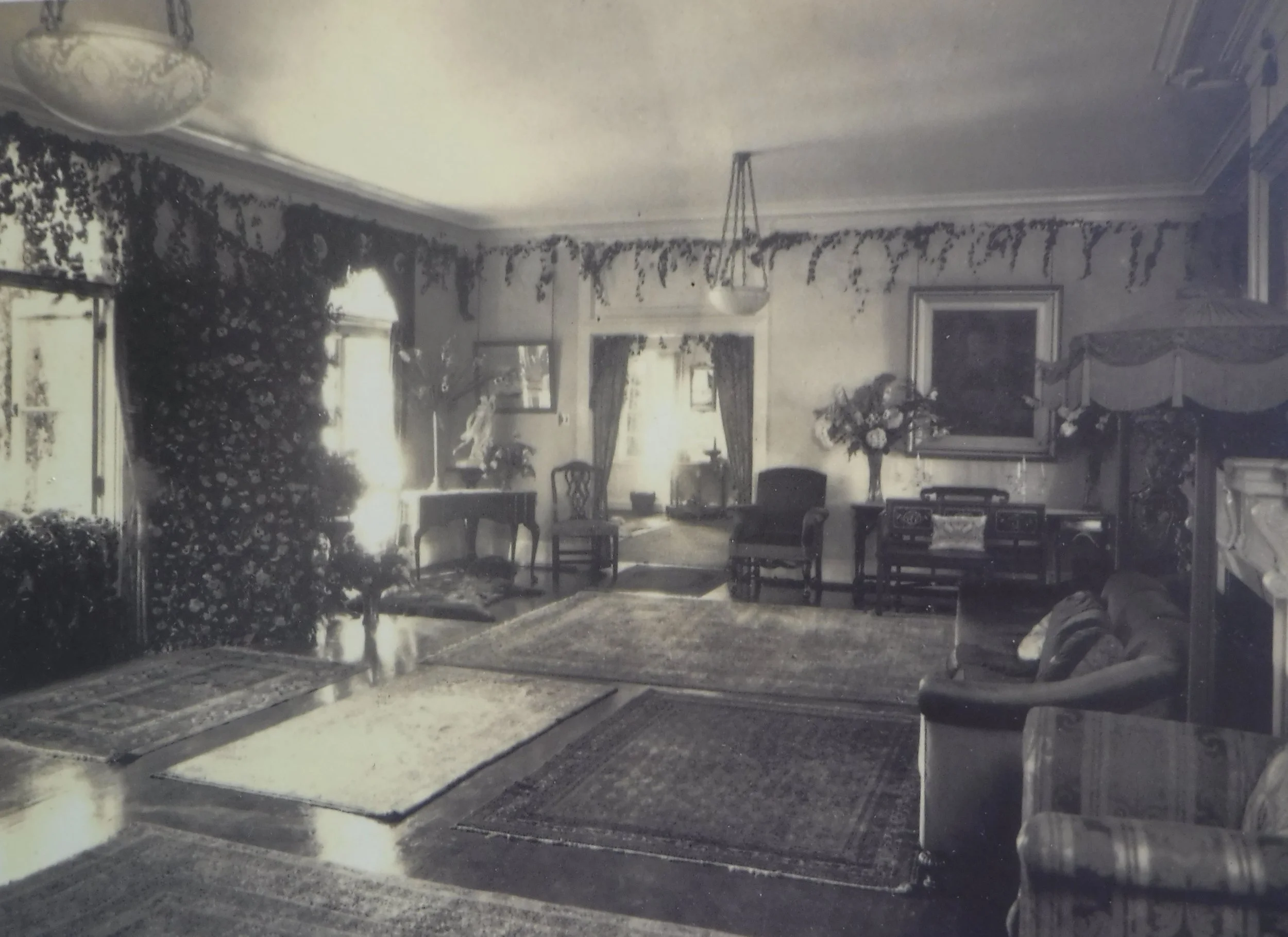 Living room with furniture and decorations, including a floral arrangement on a table, portraits on the wall, and drapes on the windows, shown in a vintage photograph.
