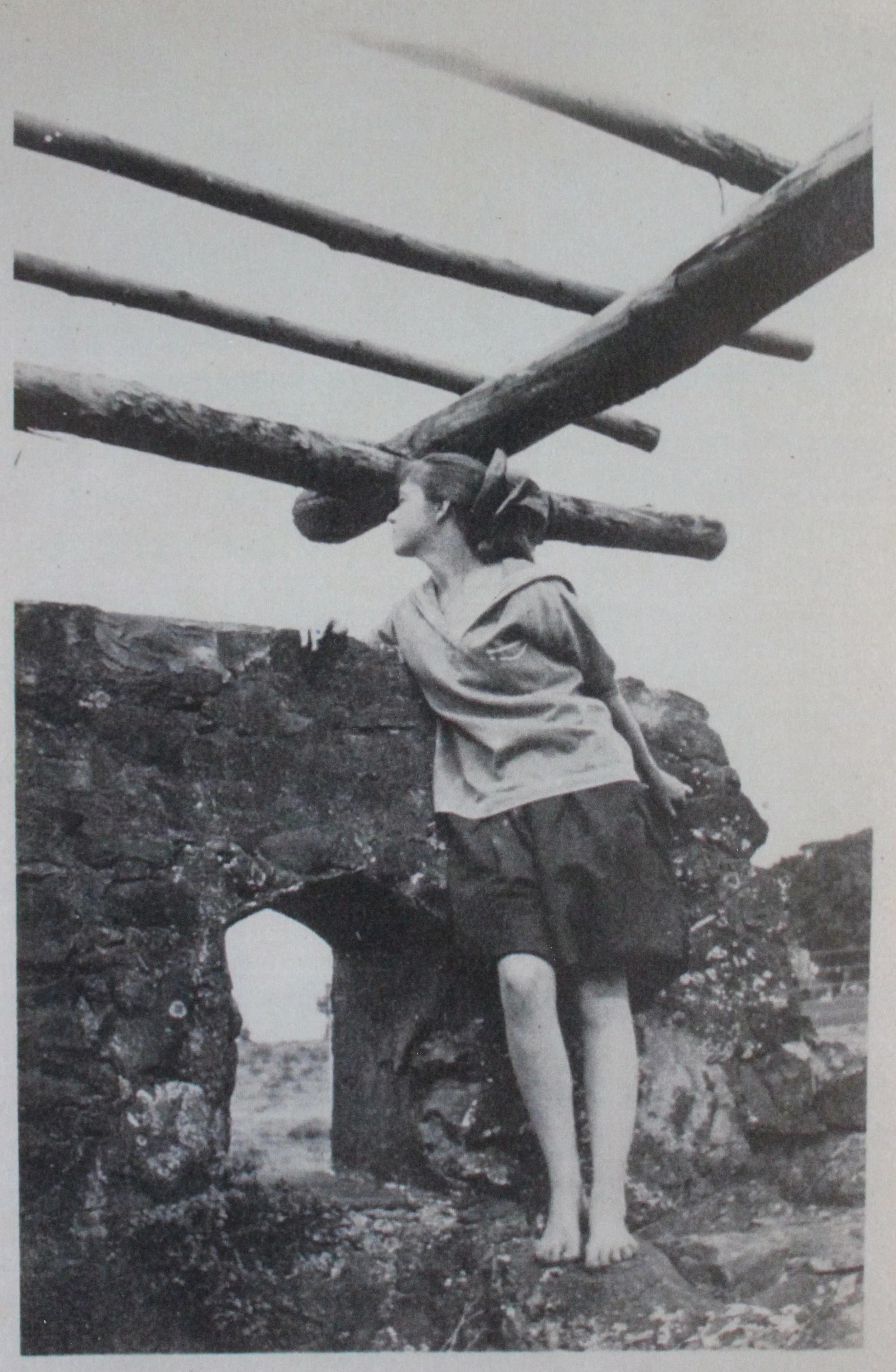 A black and white photo of a young person with a sailor hat, standing on a rock under a wooden structure, outside in a rocky area.
